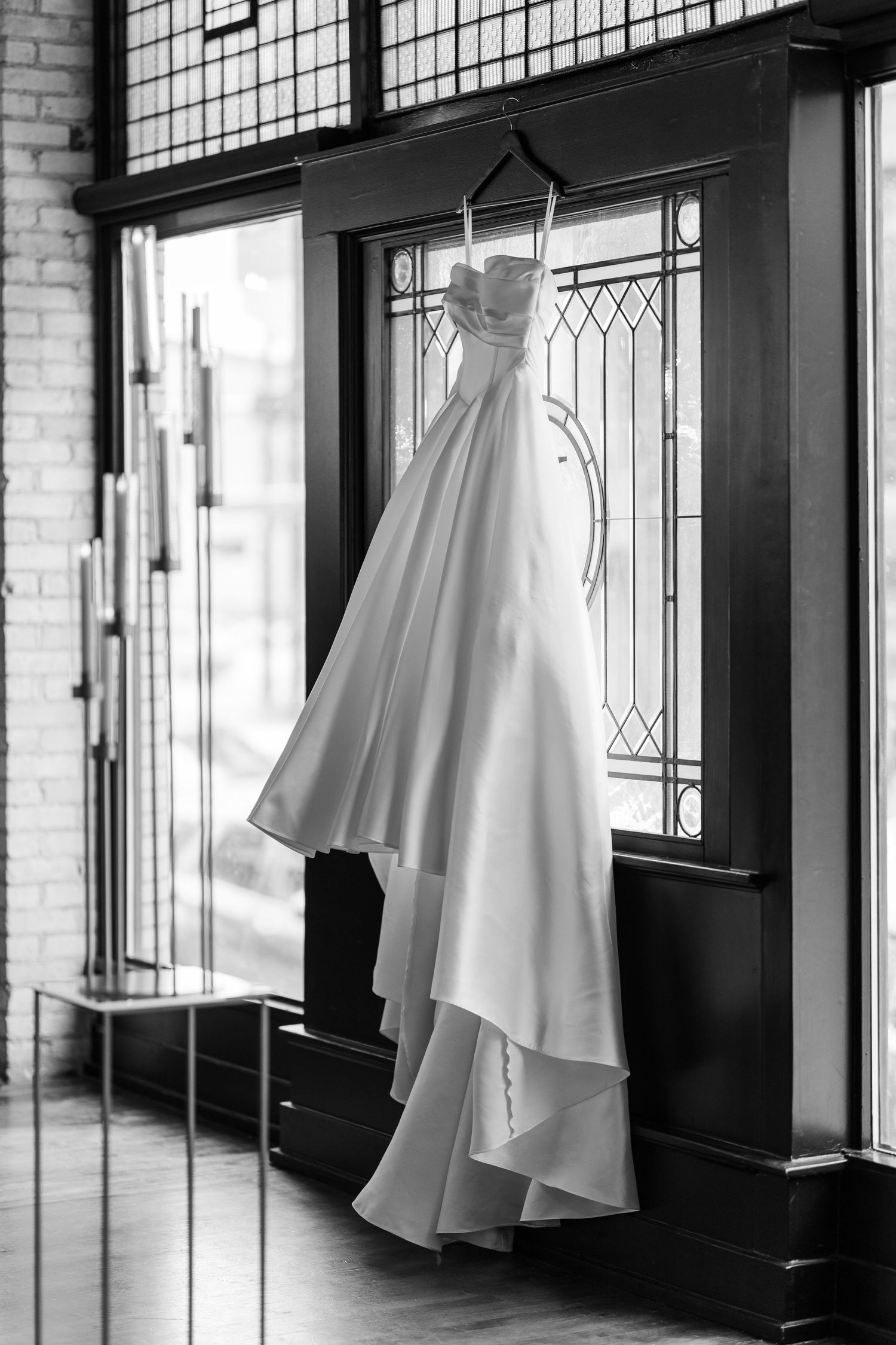 Black and white photo of an elegant strapless wedding gown hanging on a door in a vintage-style room with leaded glass windows.