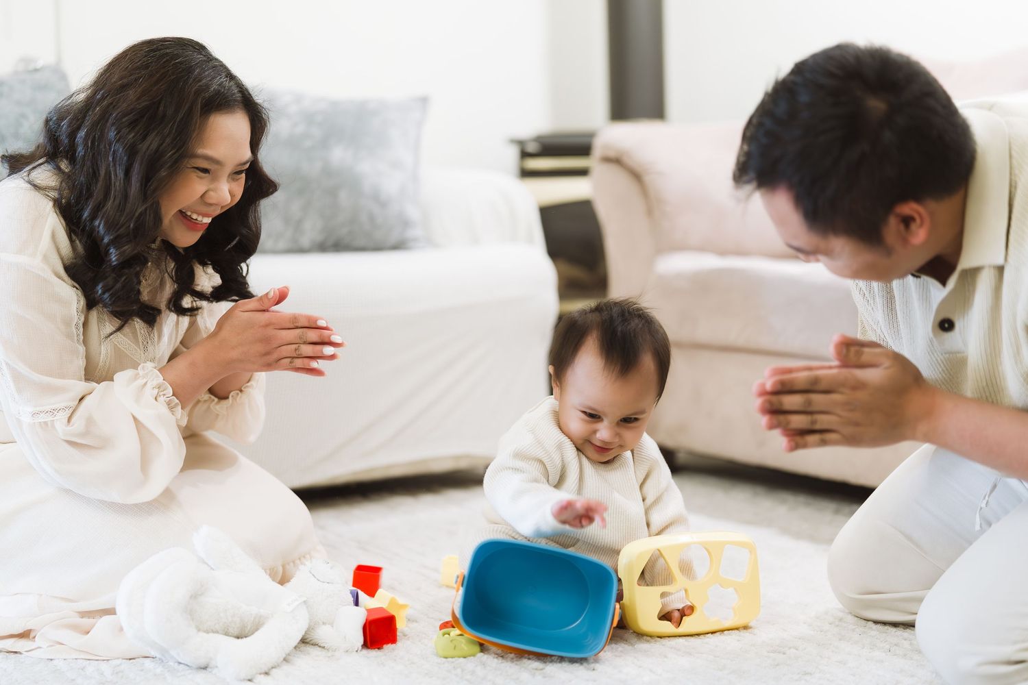 Lifestyle family photography in Victoria, BC, featuring happy parents clapping and playing with their baby on a cozy living room rug.