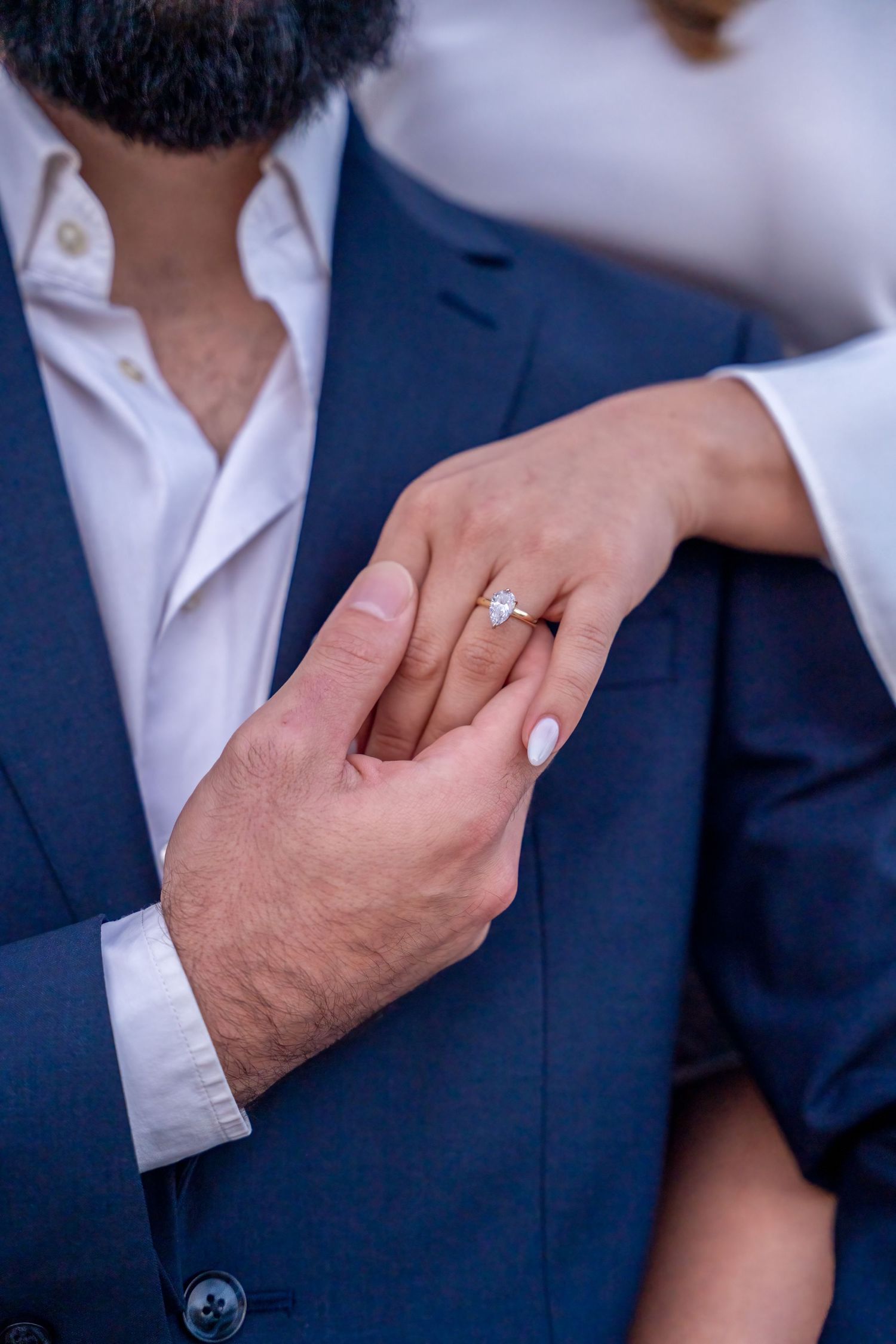 Close-up of a woman's hand with an engagement ring resting on her partner's suit, simple and elegant.