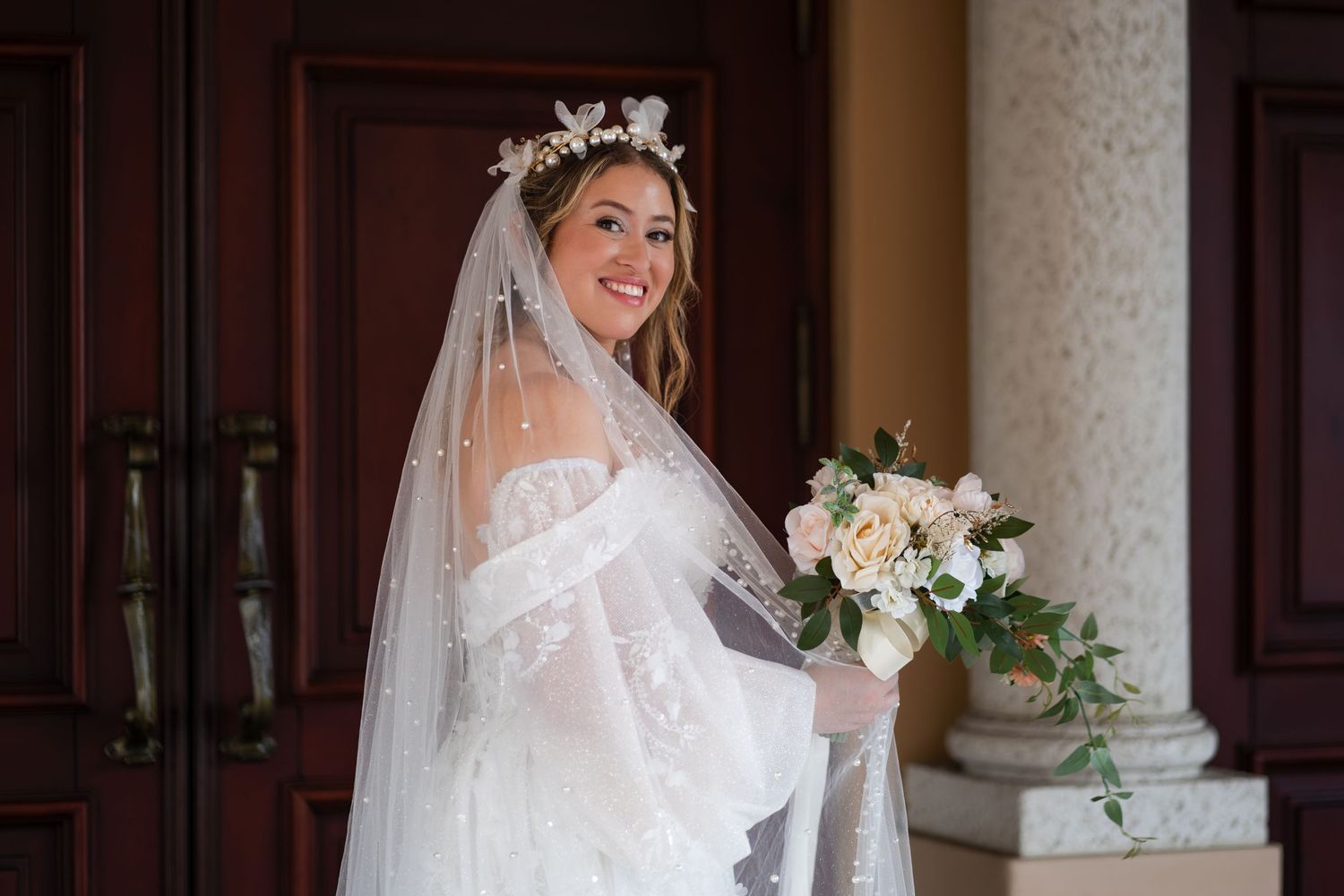 Bride in romantic off-shoulder lace wedding dress holds a blush and white bouquet while standing by wooden doors.