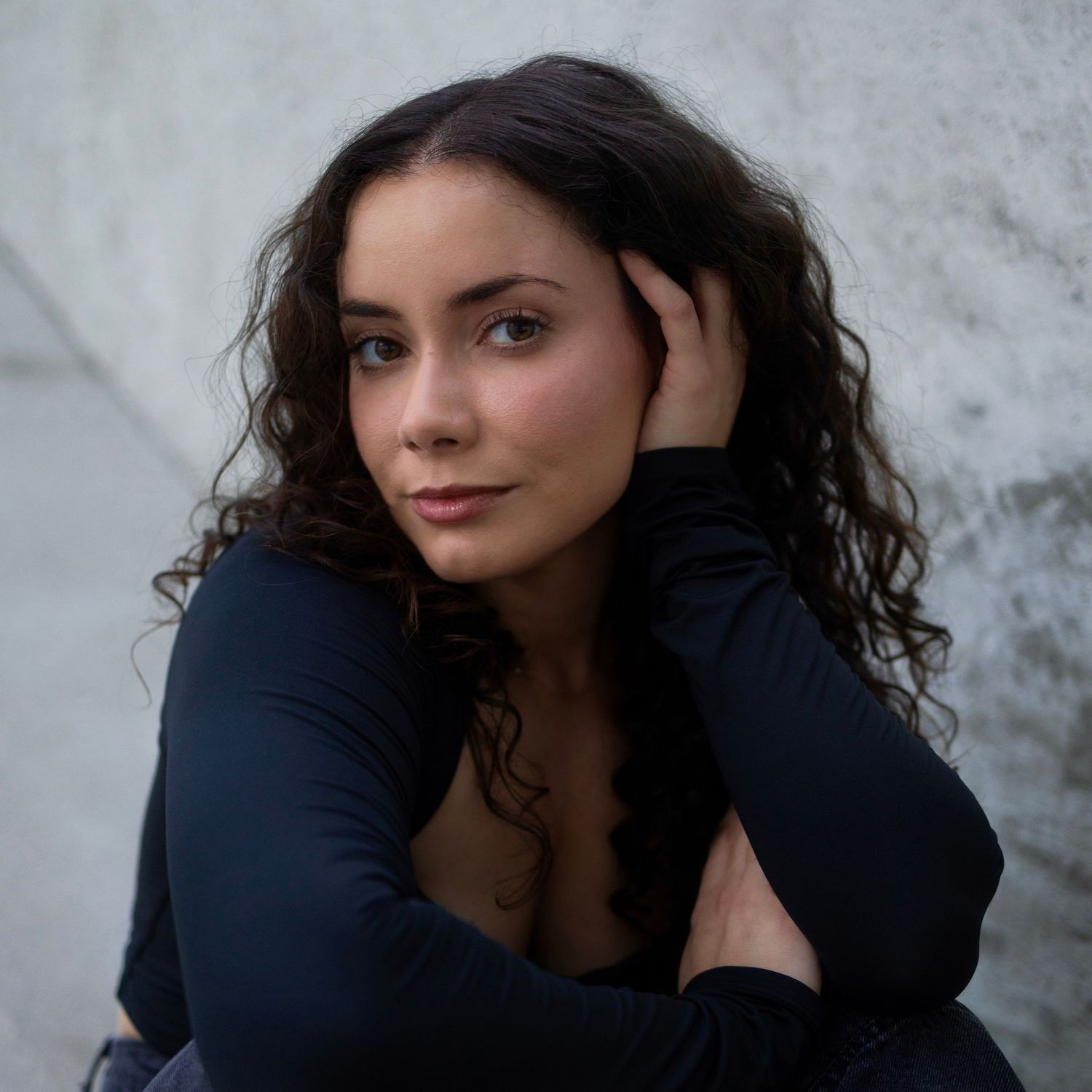 Natural light portrait of Nakita with curly hair, posing confidently against a light concrete background.