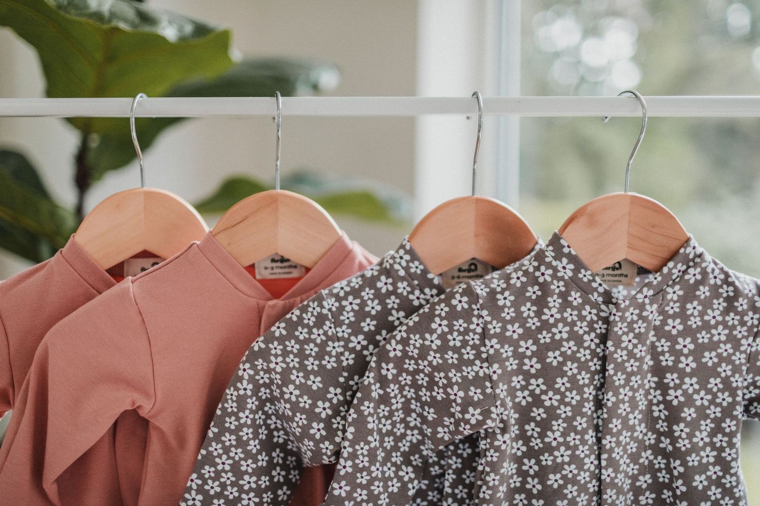 Close up image of pink baby rompers and purple flower patterned baby rompers hanging in front of a window.