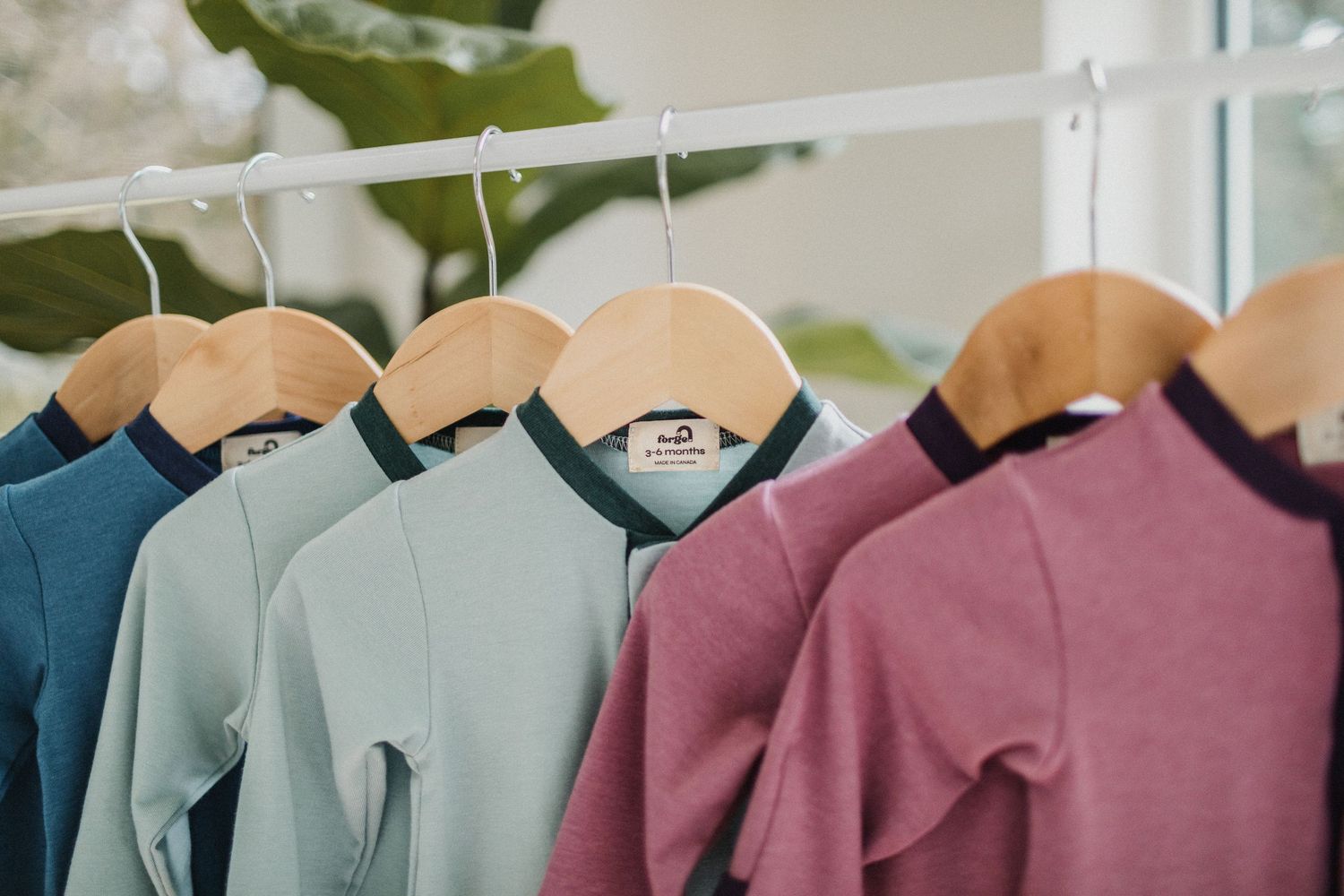 The top half of three blue, aqua, and purple baby rompers hanging in front of windows with greenery in the background.