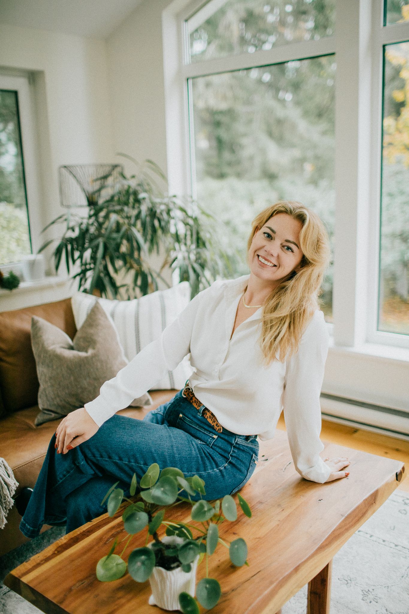 Image of a woman comfortably sitting on a table smiling at the camera. She is surrounded by plants.