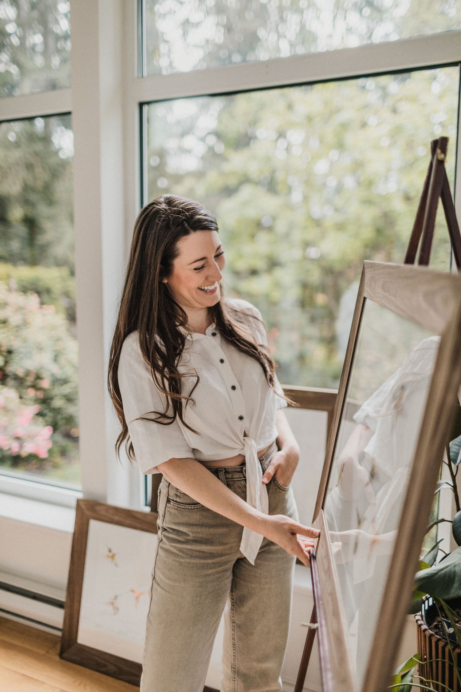 A female artist laughing while touching and looking at her painting. Windows, trees and another painting are in the background.