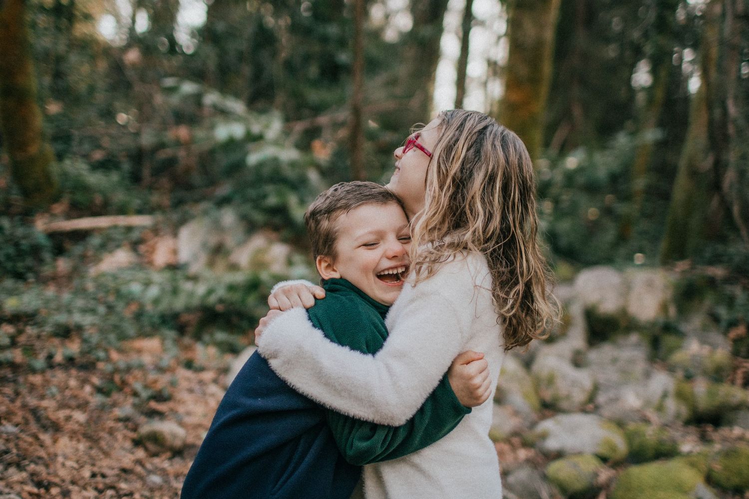 A five-year-old brother and eight-year-old sister, hugging and laughing with trees, rocks, and blue sky in the background.