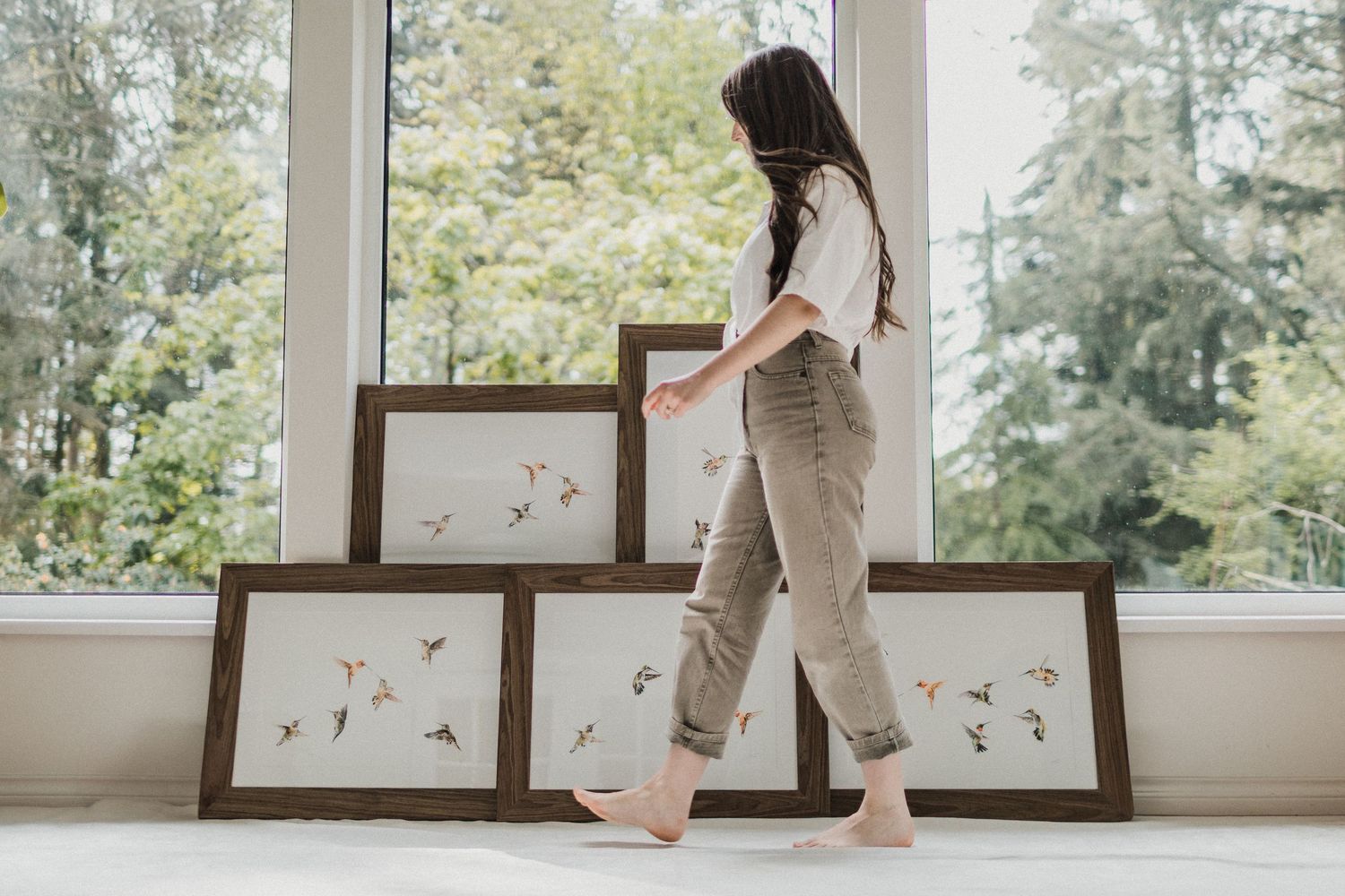 A female artist walking in front of five watercolour paintings of humming birds. There are large windows and trees in the background,
