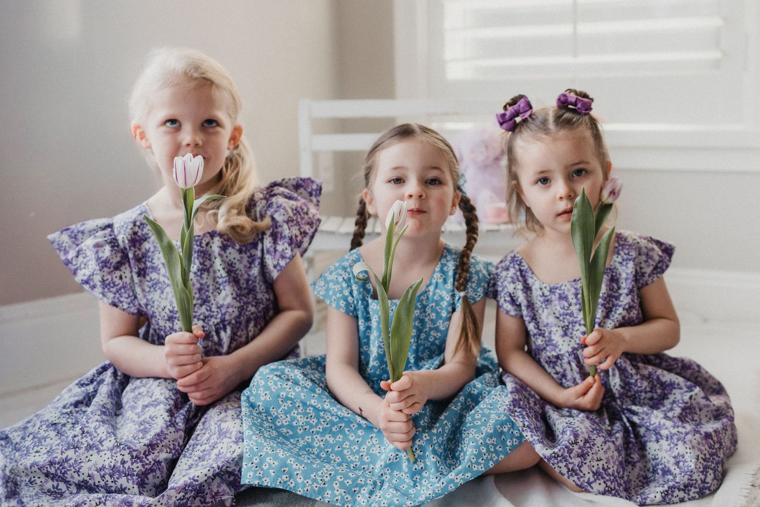 Three young girls sitting in a row on the ground holding tulips wearing purple and blue dresses.
