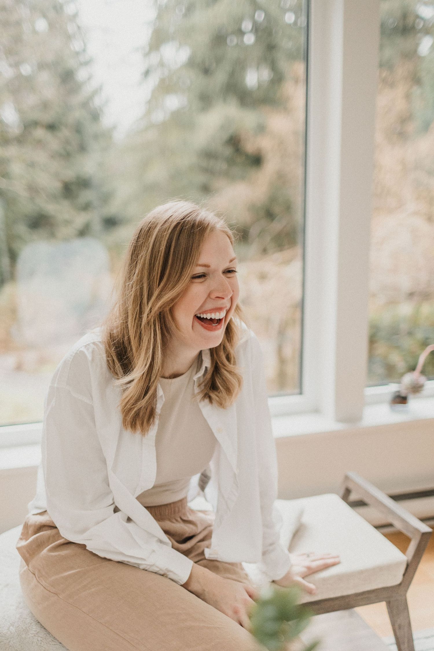 Woman sitting with hands on lap laughing. Large windows and trees are in the background.