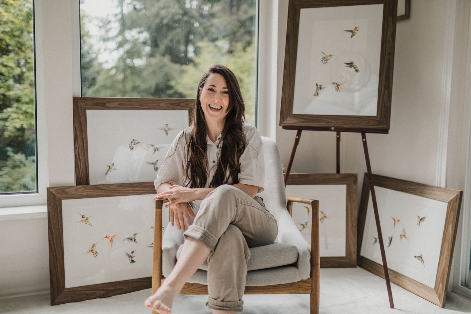A female artist smiling and sitting in a chair in front of five humming bird watercolour paintings.