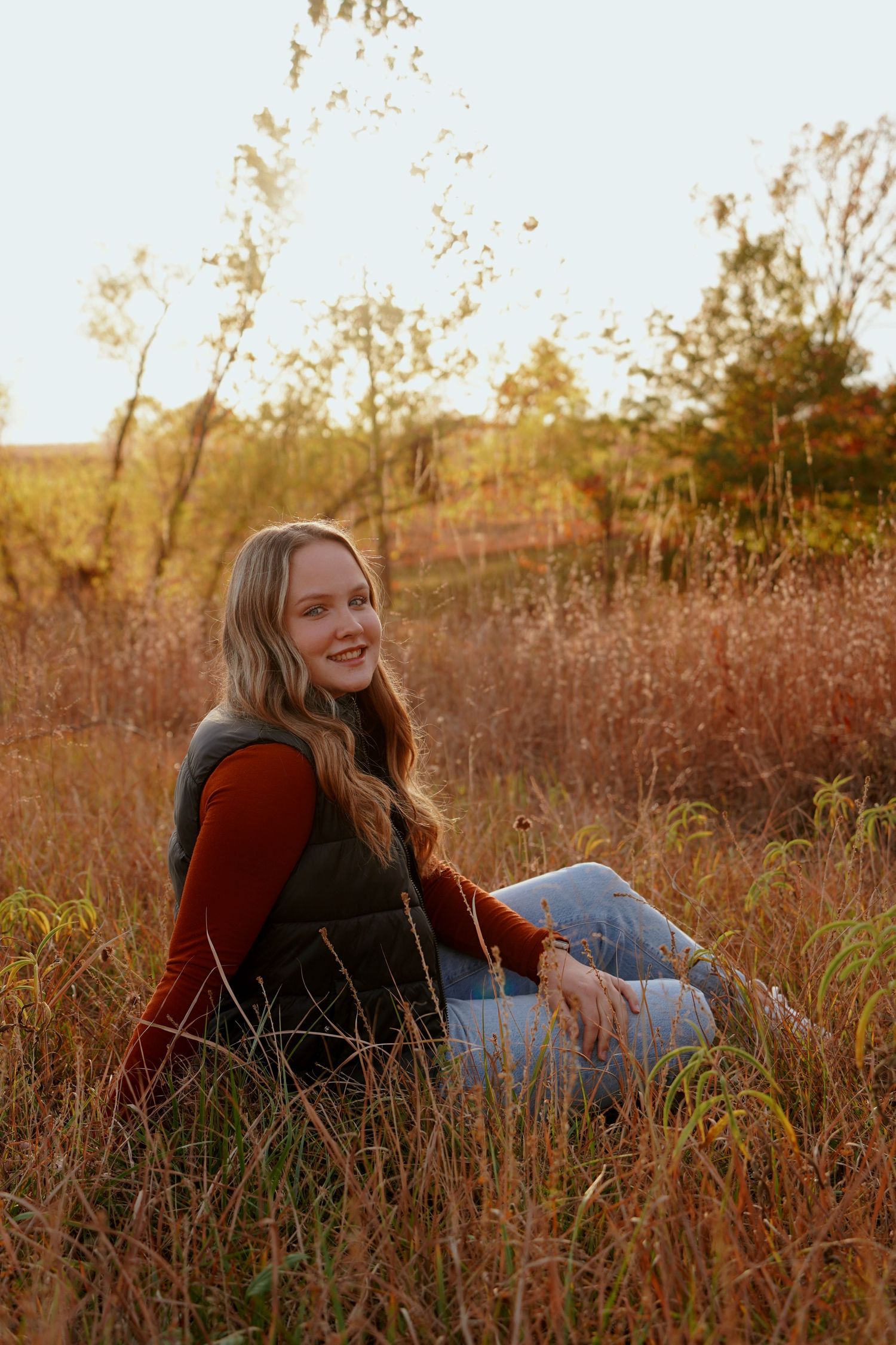 Blonde girl in rust top, vest, and jeans sitting in golden autumn meadow grass, smiling softly.
