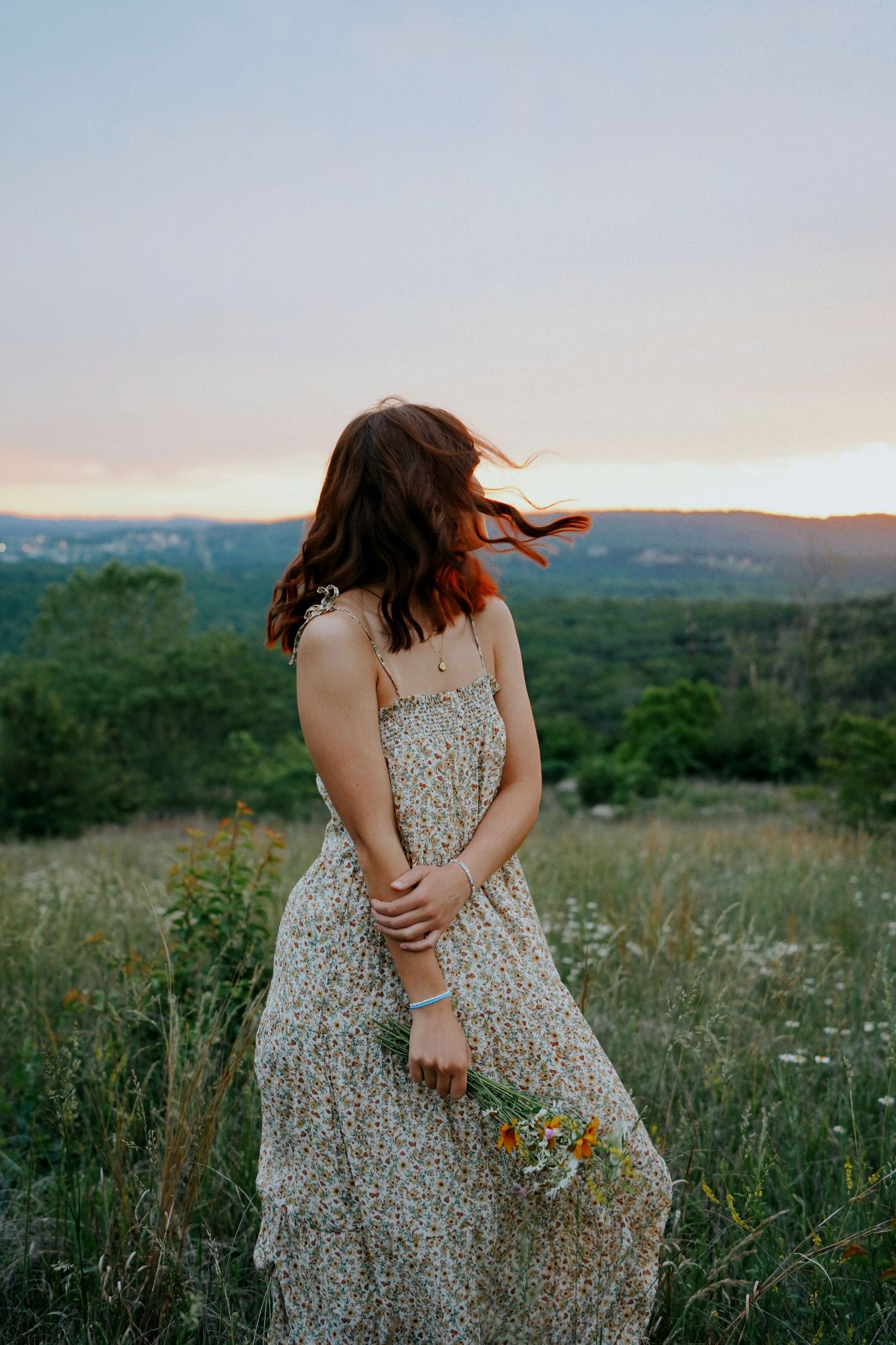 A young woman in a floral sundress holds wildflowers while her hair blows in the wind at sunset.