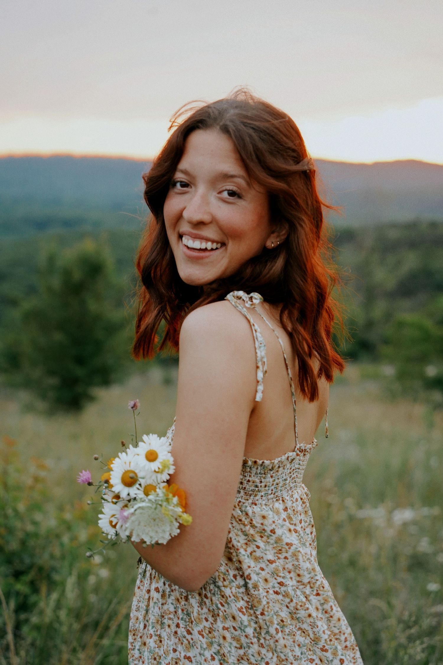 A smiling young woman with auburn hair holds daisies while posing in a lush green field at sunset.