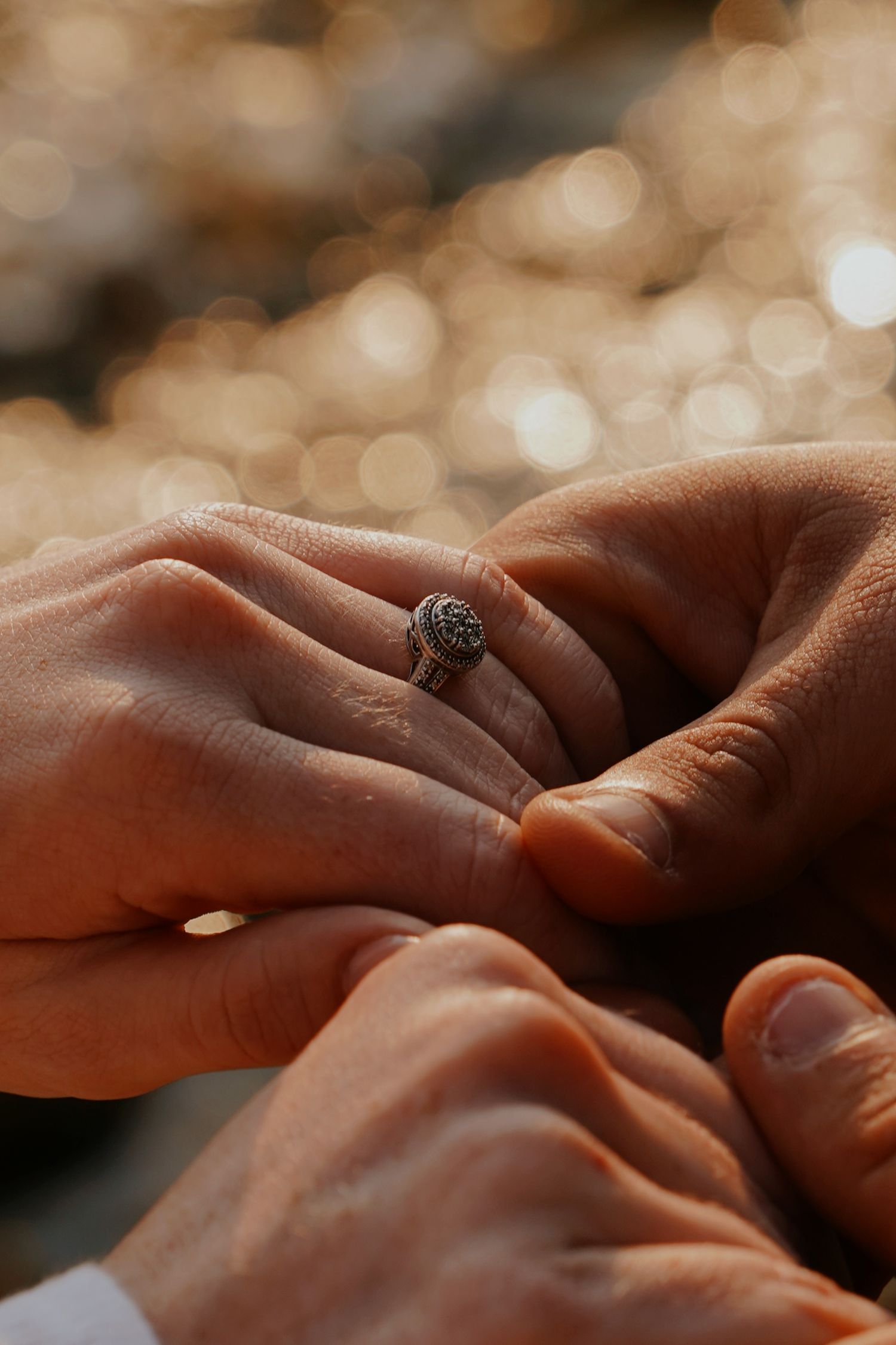 Close-up of hands with ornate engagement ring in warm bokeh light.