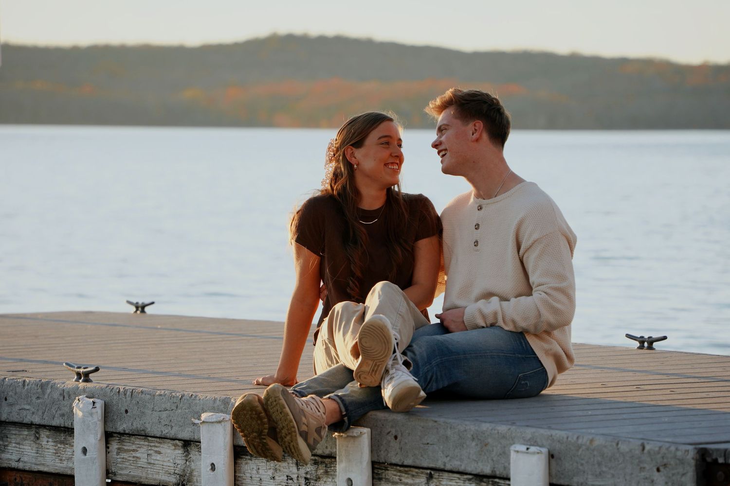 A happy couple sits together on a lakeside dock enjoying a warm sunset conversation.