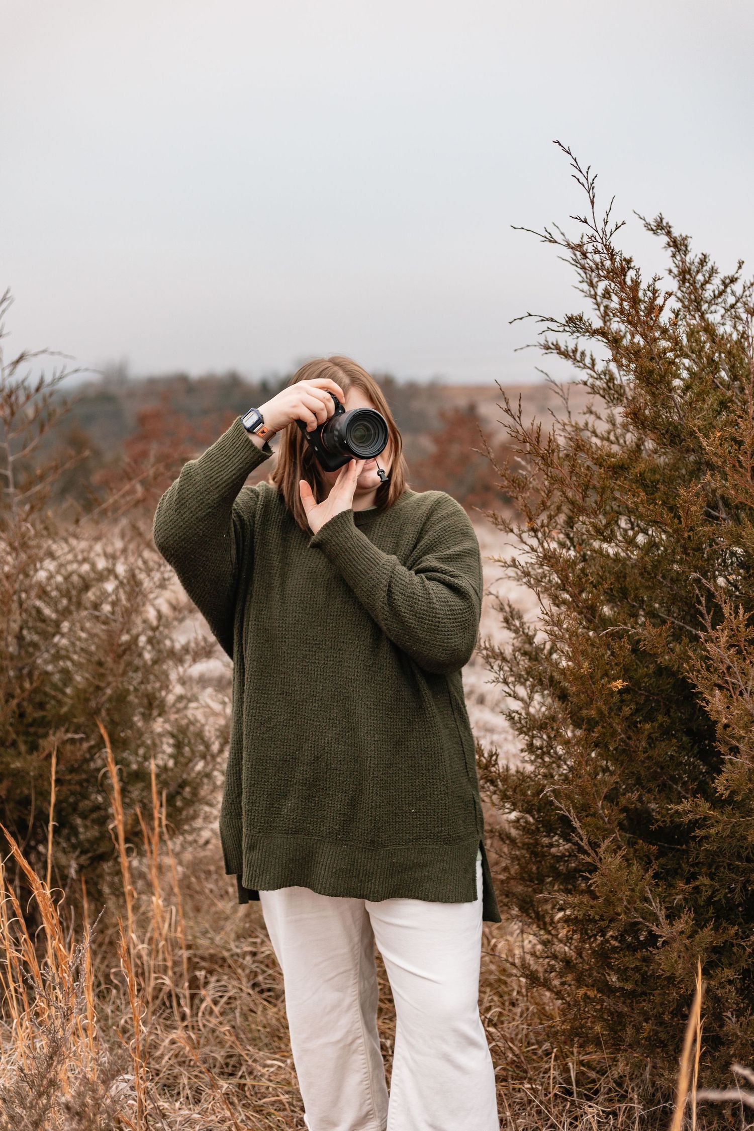 Female photographer in olive green sweater photographing with DSLR camera in a fall landscape.