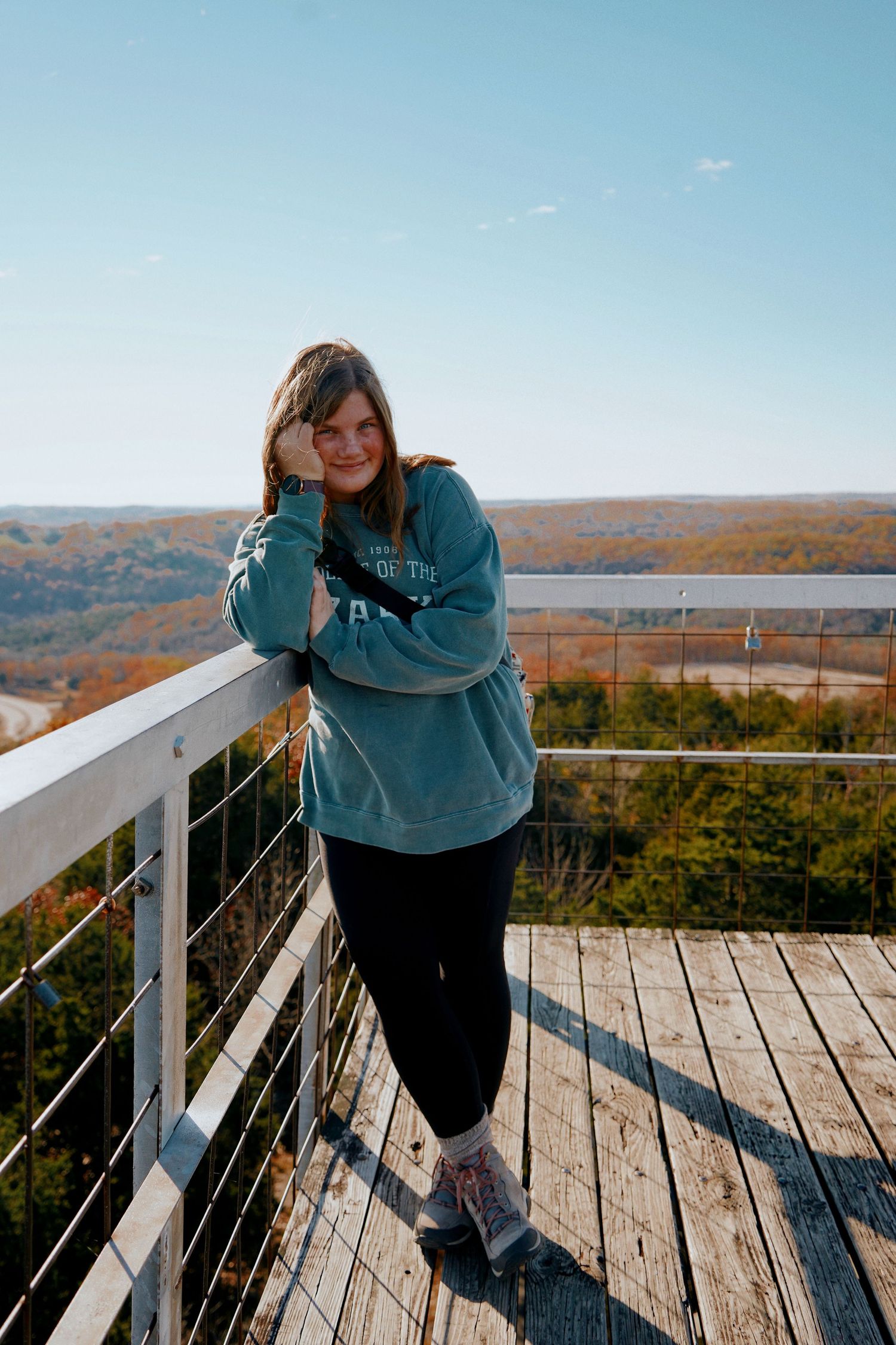Smiling woman in teal sweatshirt leaning on railing of scenic overlook with colorful autumn valley view.