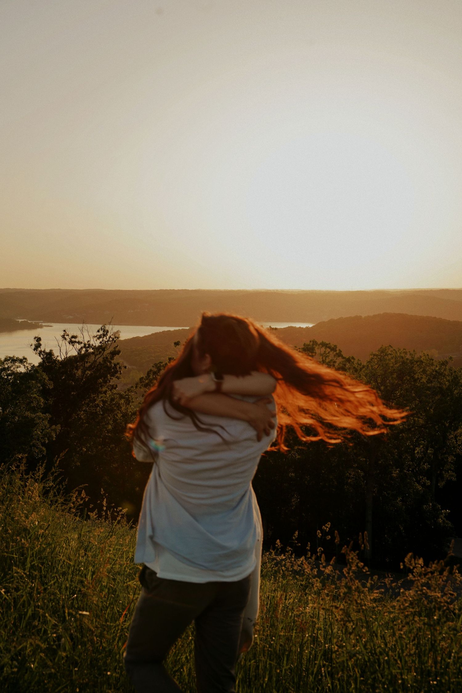 Woman with flowing red hair hugging someone at sunset overlooking a scenic river valley landscape.