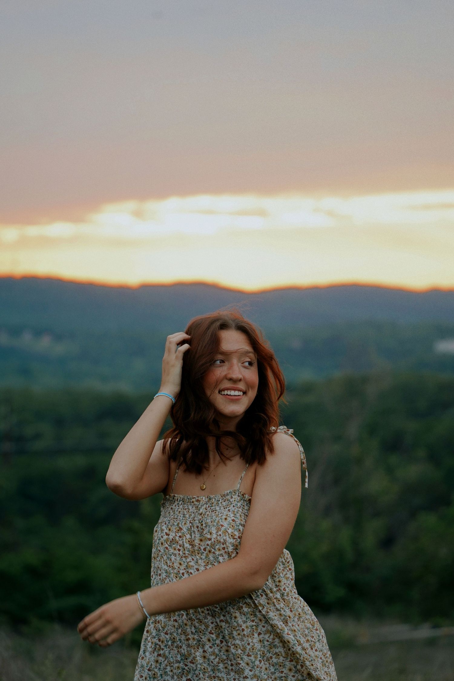A joyful young woman in a floral dress laughs against a stunning mountain sunset backdrop.