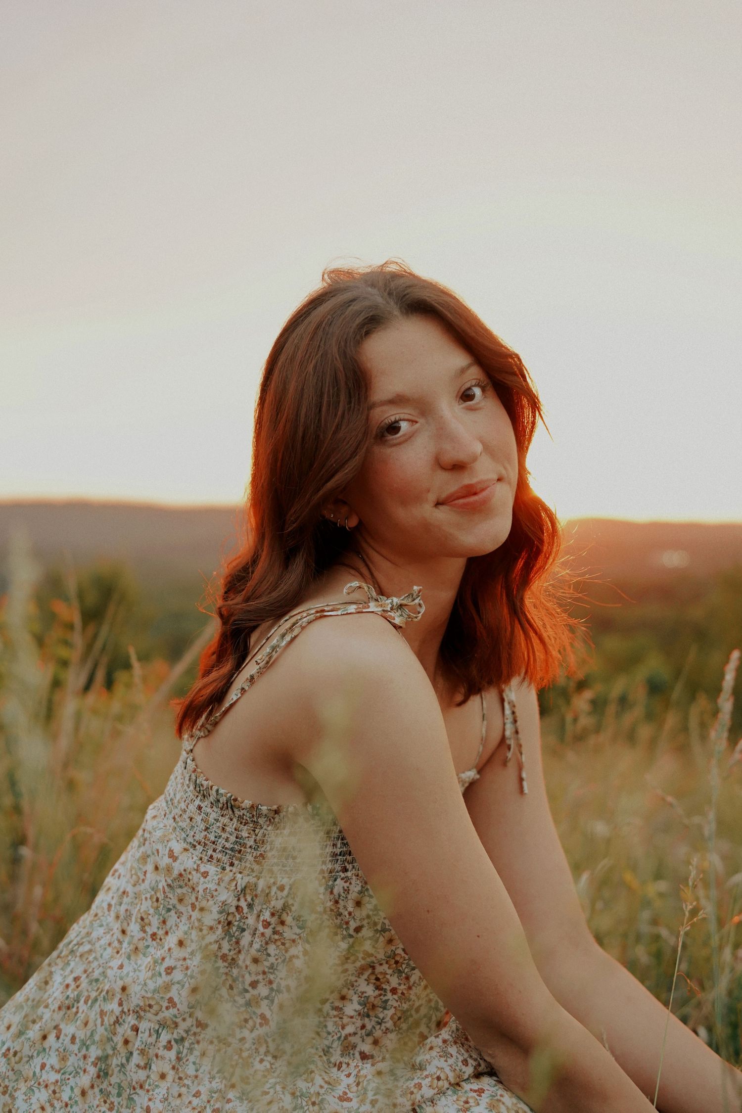 A young woman with auburn hair in a floral sundress sits in a golden field at sunset.