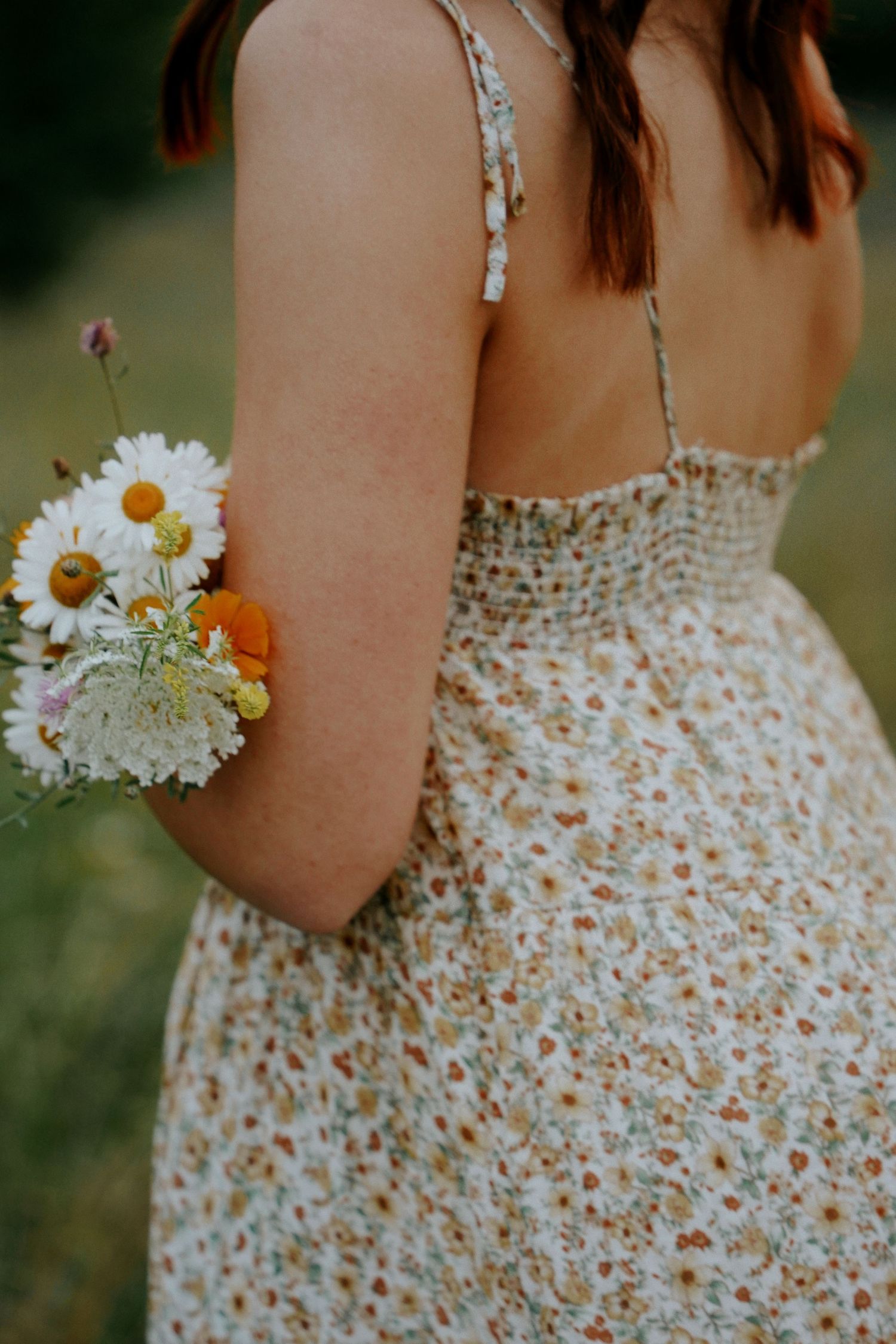 Close-up back view of a young woman in a floral sundress holding a bouquet of daisies outdoors.