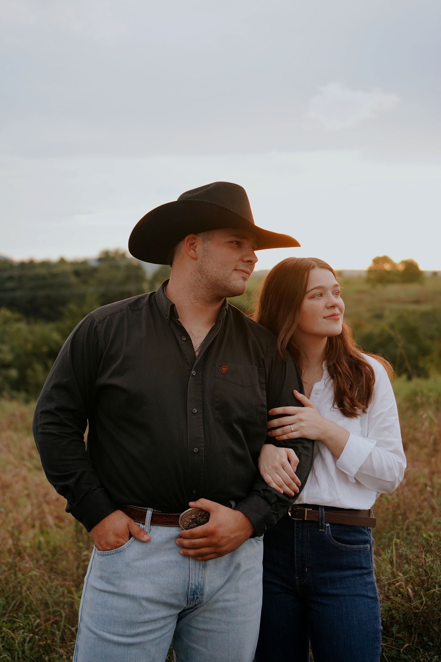 A couple poses in a golden field, man in black cowboy hat and shirt, woman in white blouse, embracing at sunset.