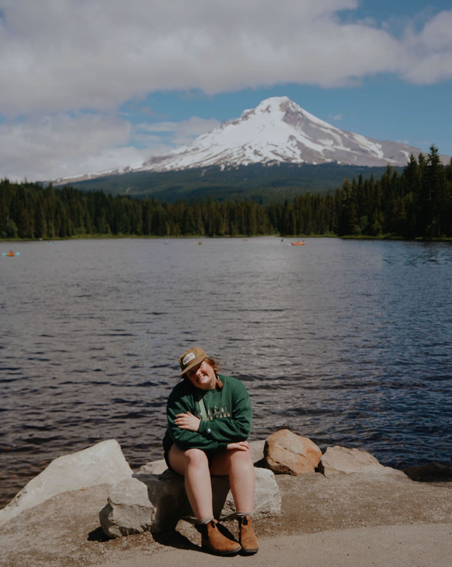 Woman in green hoodie sitting on rocks by mountain lake with snow-capped Mount Hood in background.