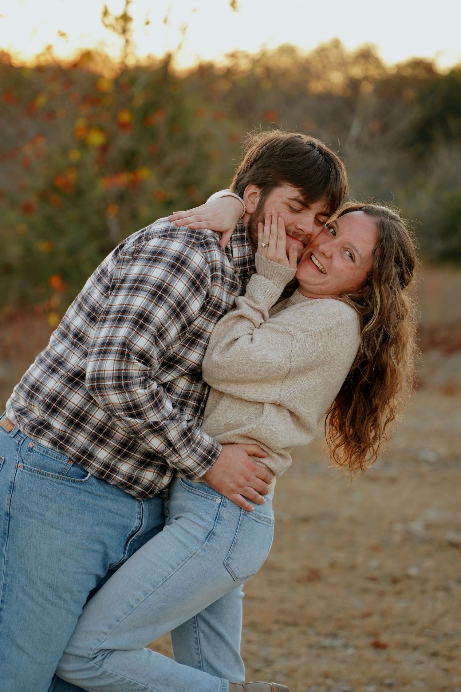 Couple embracing outdoors at golden hour, man kissing woman's cheek as she smiles, wearing flannel and cream sweater.