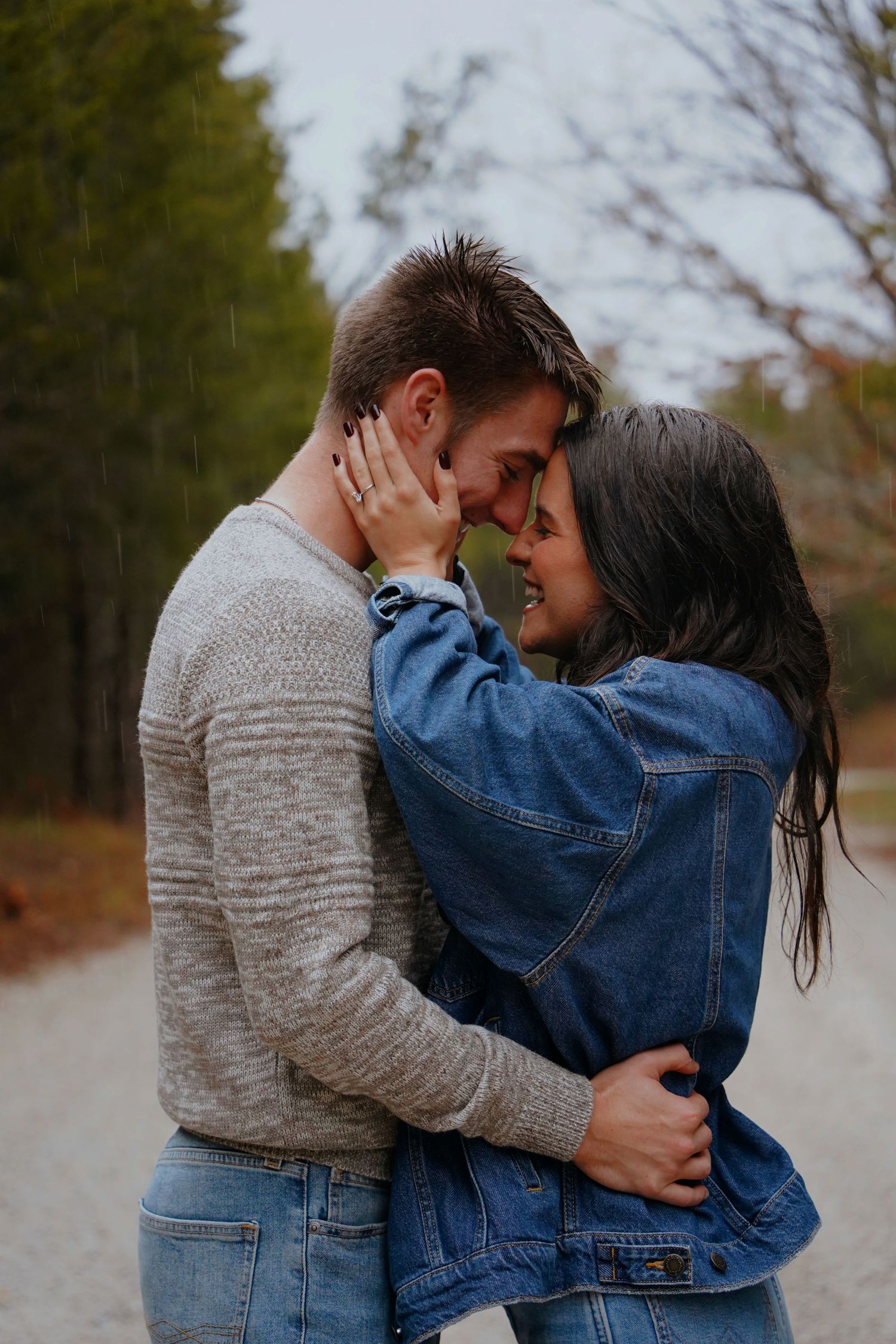 Couple embracing and almost kissing in the rain on a forest path, wearing casual jeans and sweater outfits.