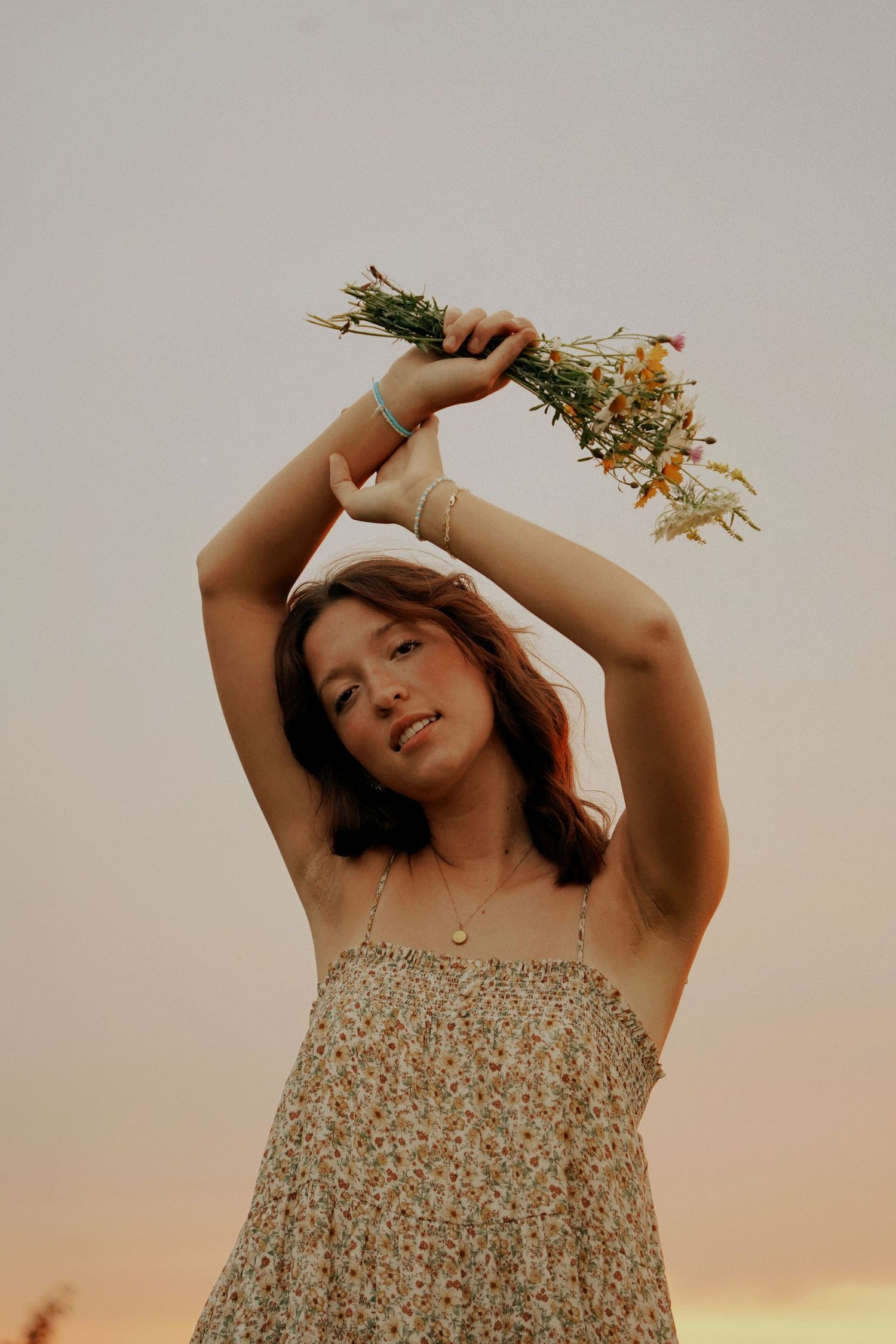 A young woman in a floral sundress raises a wildflower bouquet overhead against a golden sunset sky.