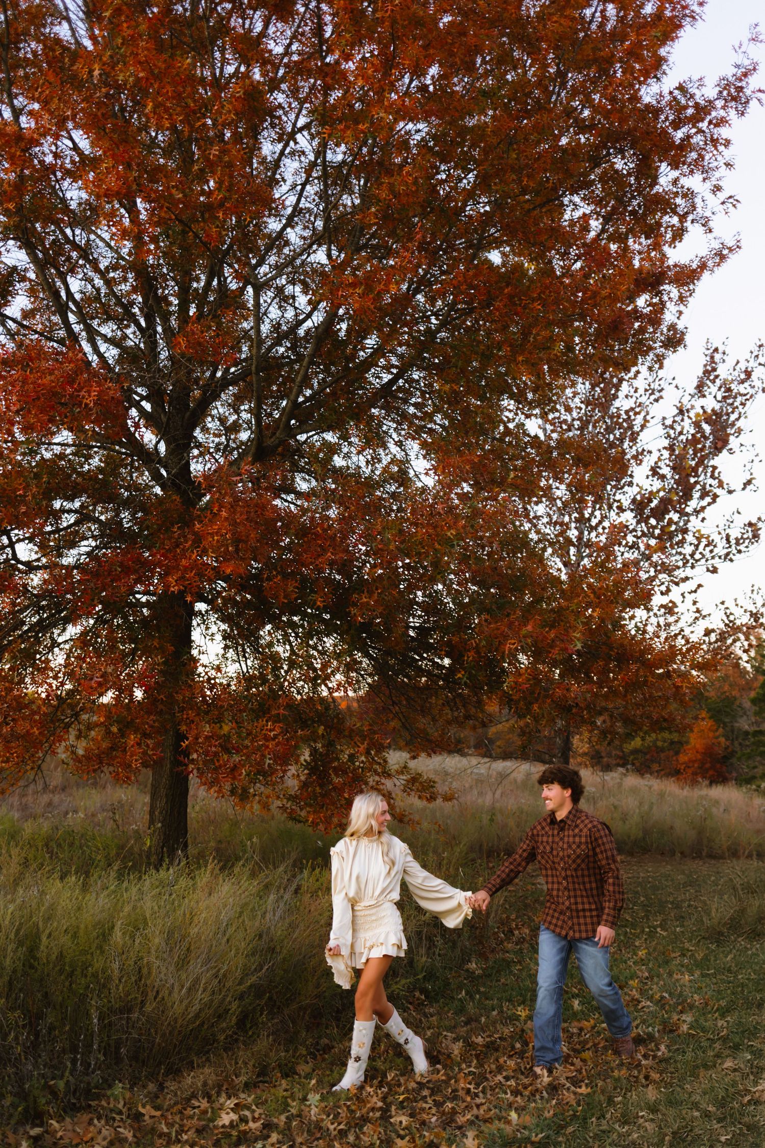 A couple holds hands beneath a large autumn oak tree with vibrant orange leaves in a grassy meadow at sunset.