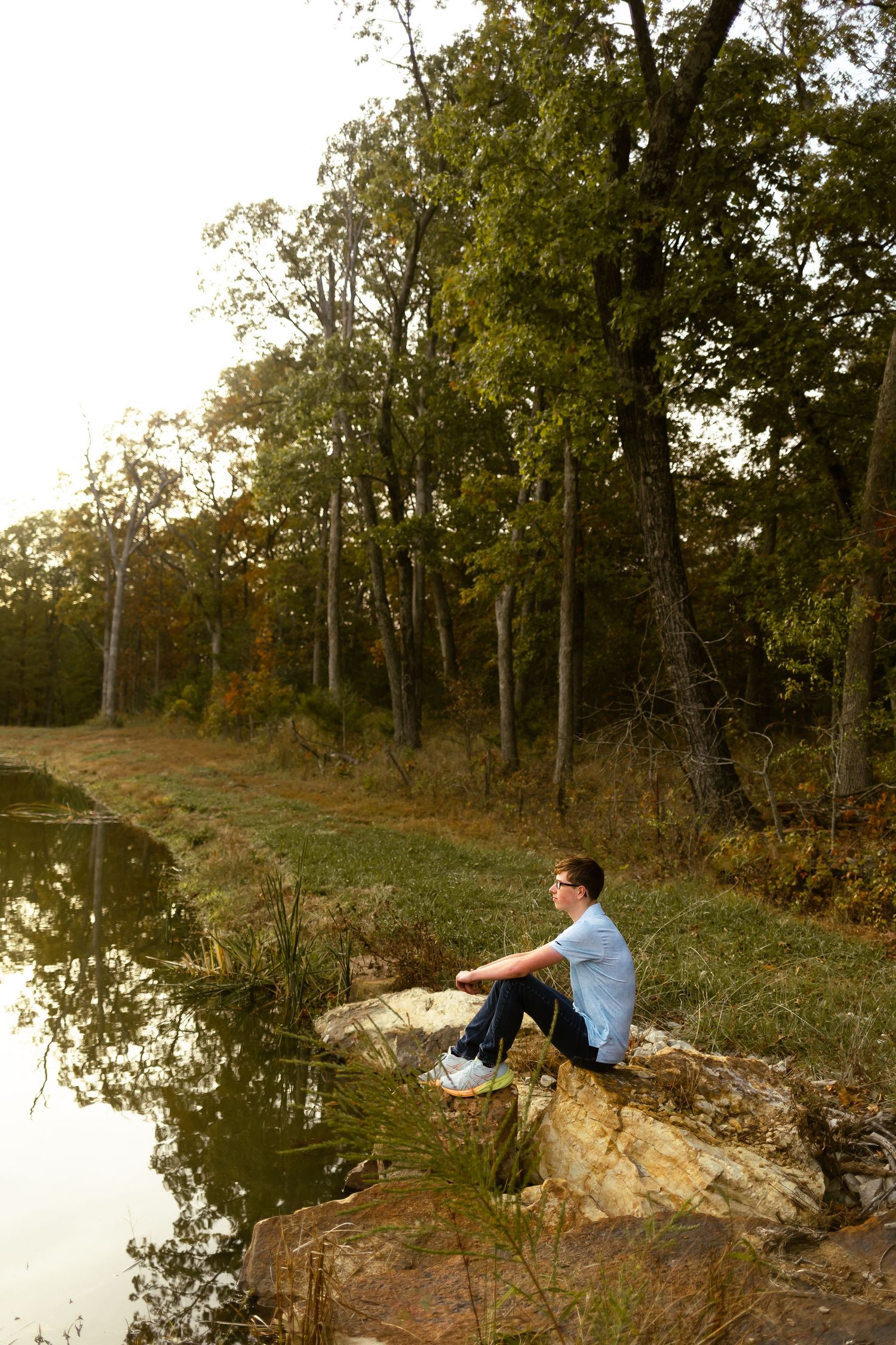 A person sits peacefully on a rocky outcrop overlooking a tranquil lake surrounded by autumn trees at sunset.