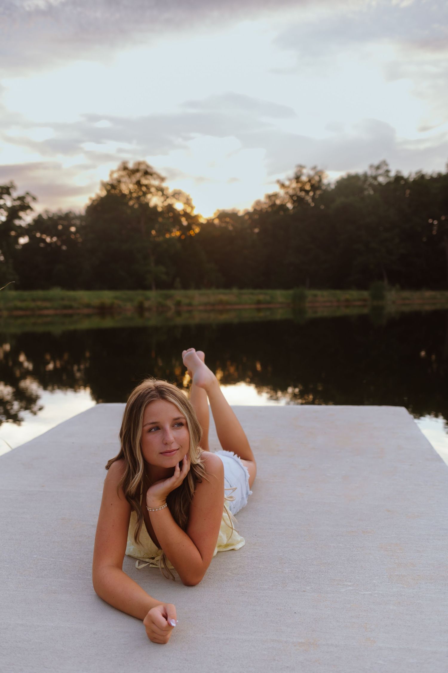Someone lying on a white surface near a tranquil lake at sunset with trees in the background.