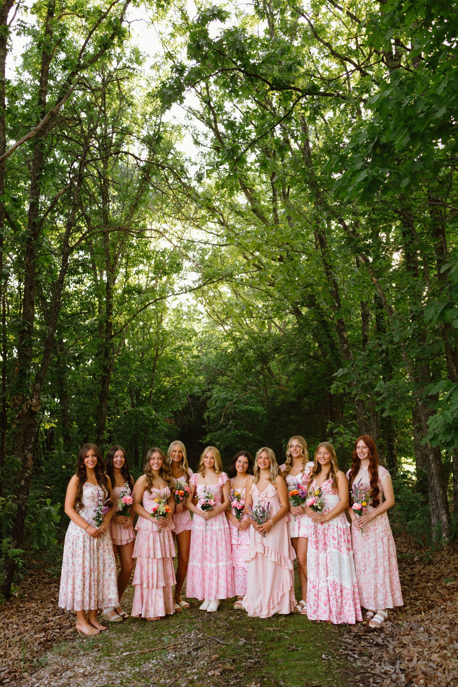 Bridesmaids in pink floral dresses holding bouquets pose together in a lush green forest setting.