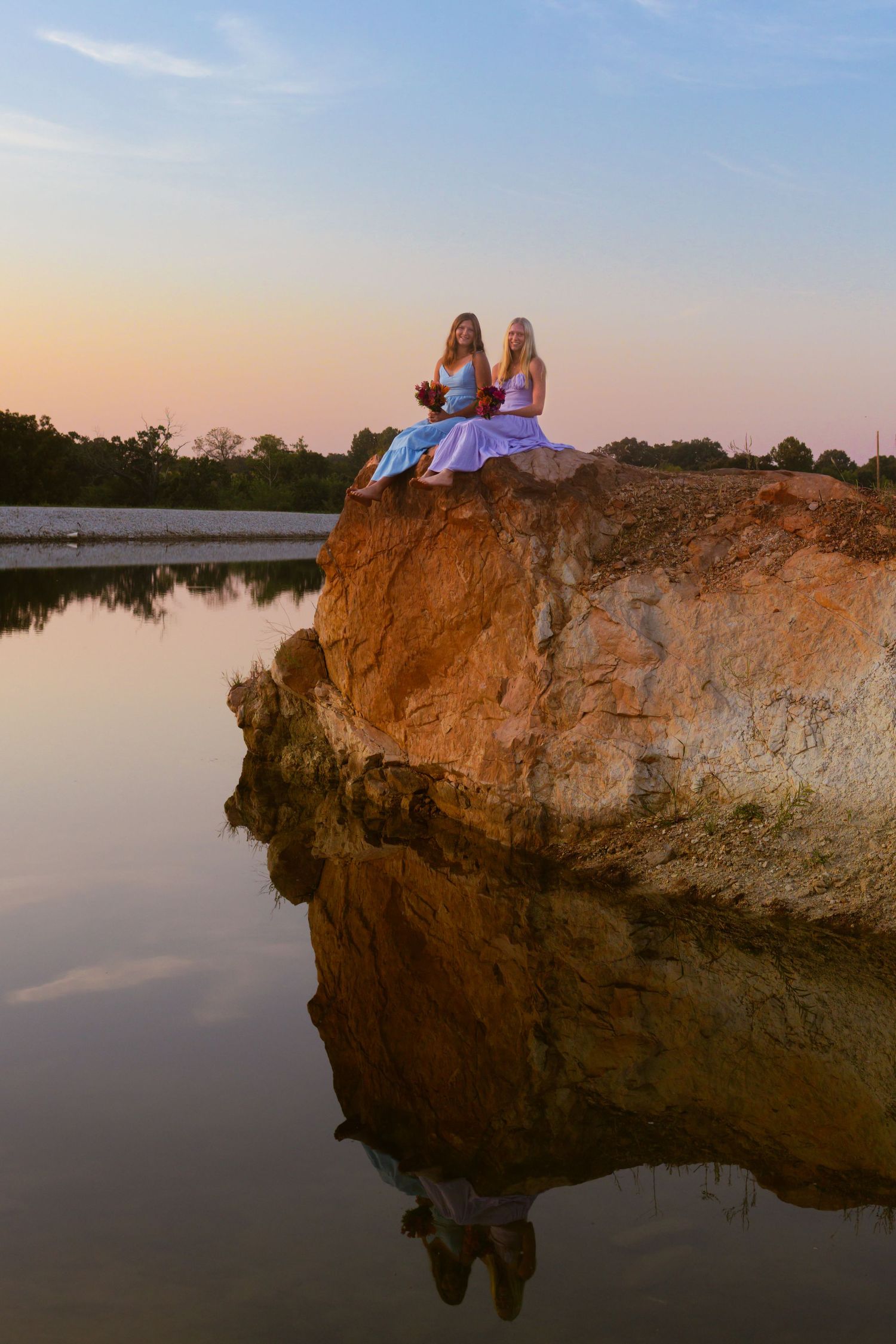 Two figures sit atop a rocky cliff overlooking a tranquil lake at sunset while their reflection appears in the calm water below.