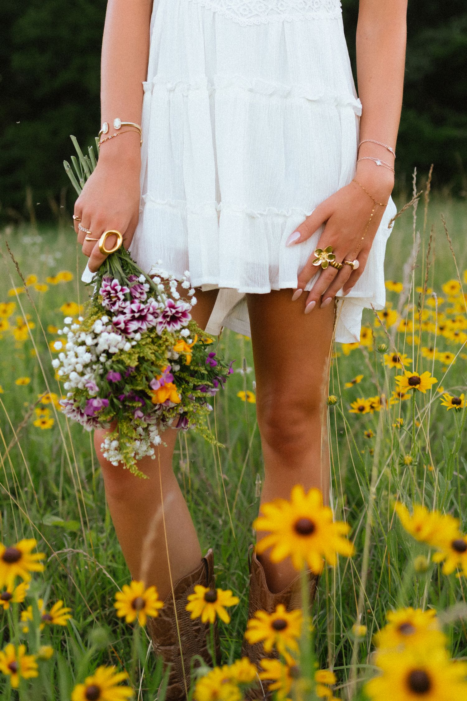 Woman in white dress and cowboy boots holding wildflowers in a sunflower field.