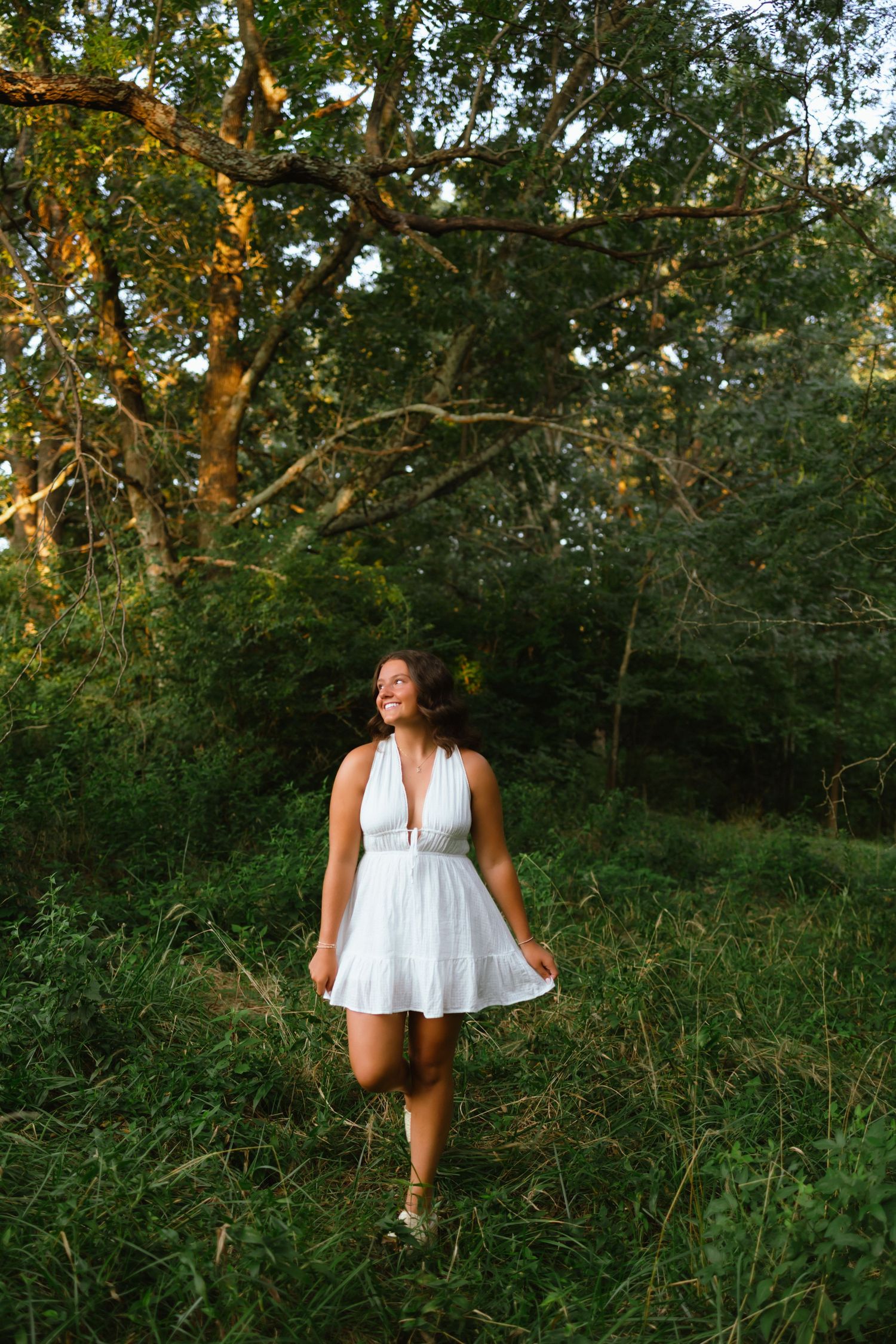 Young woman in white sundress smiling while walking through lush green forest during golden hour.