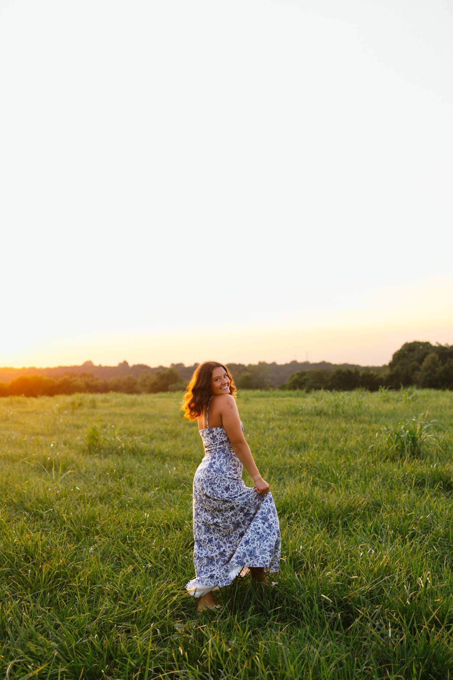 Woman in floral maxi dress twirling in green field at golden sunset, smiling over shoulder.