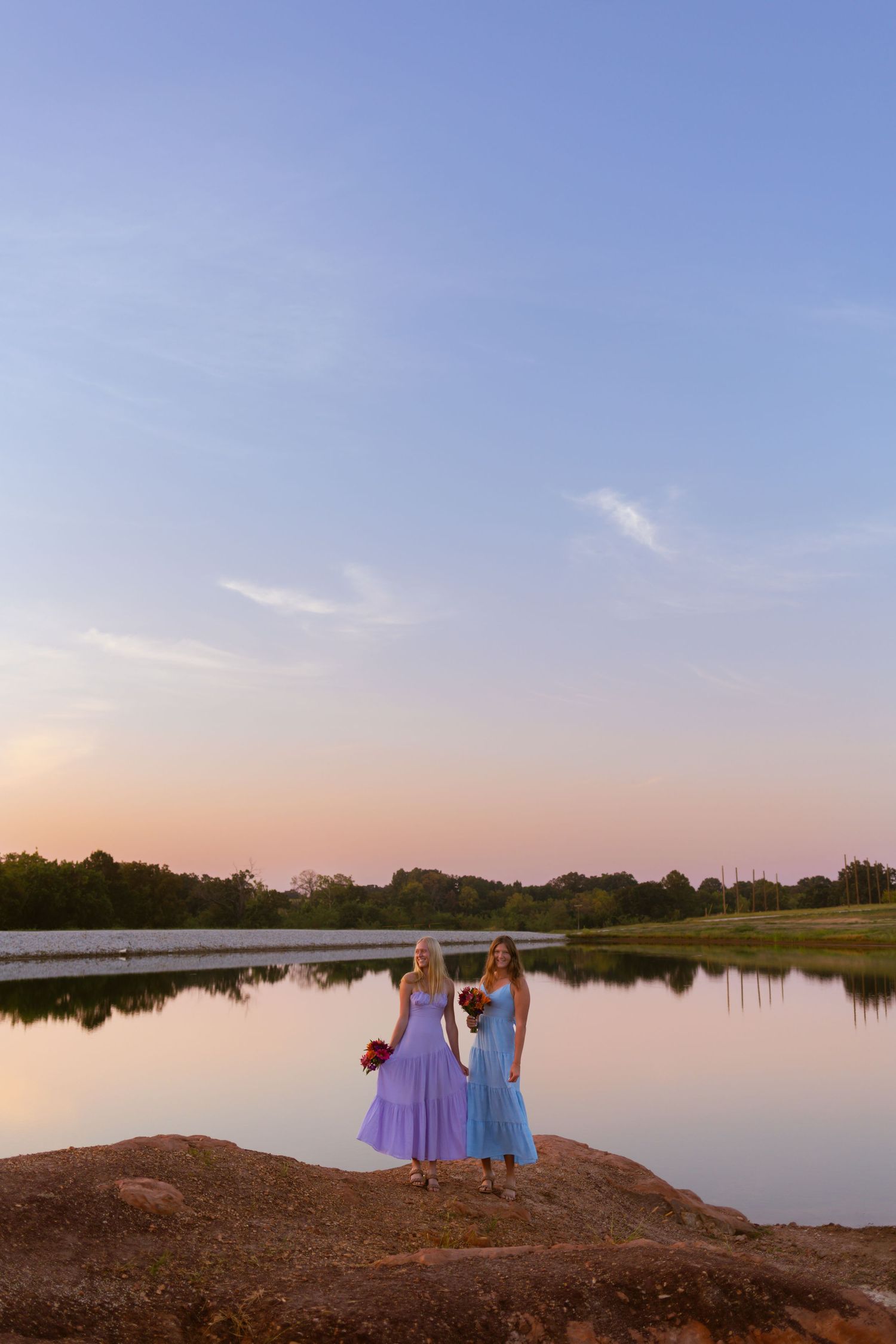 Two figures in purple and blue dresses stand by a serene lake at sunset as the sky fades from blue to pink.