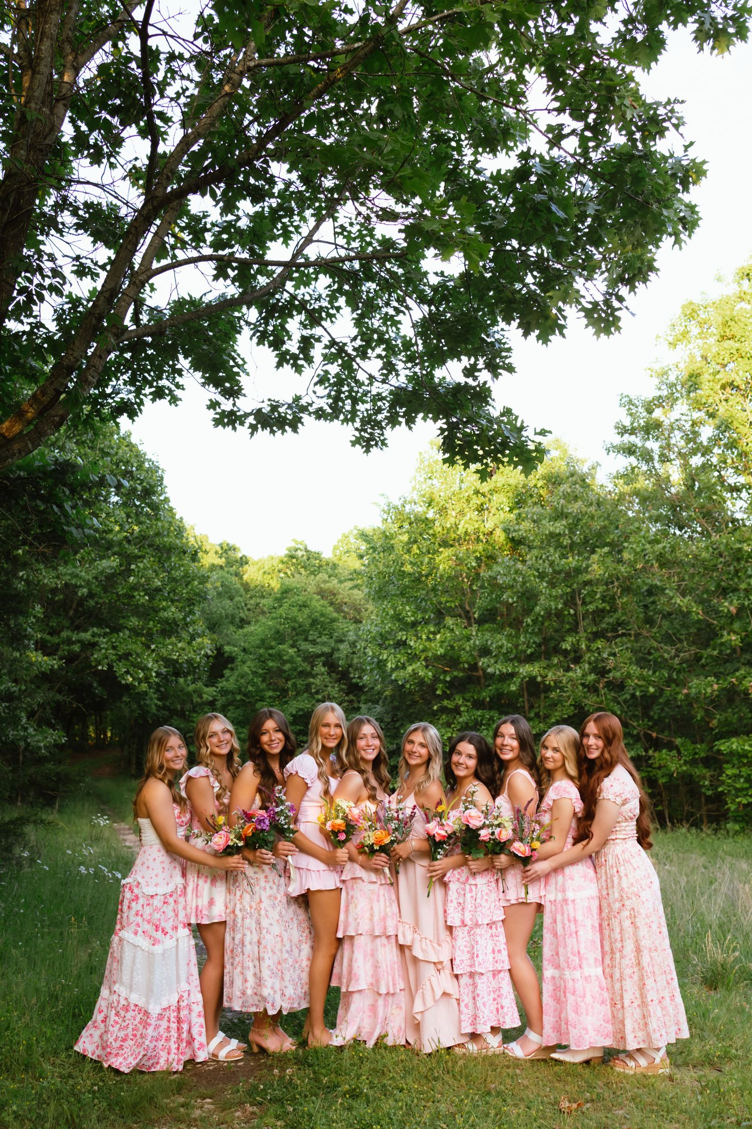 A group of bridesmaids in pink floral dresses holding bouquets, posing outdoors under a large green tree.