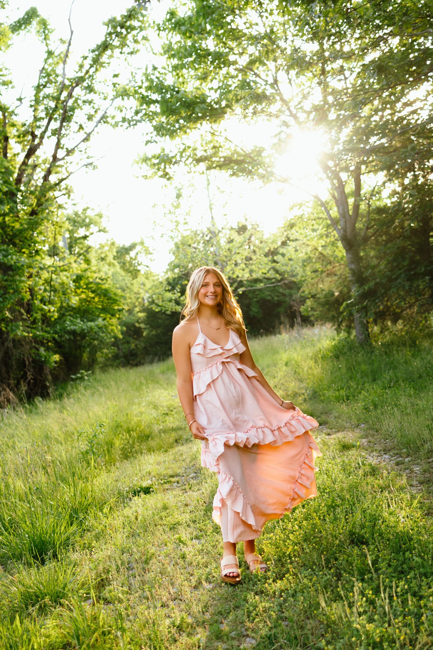 Young blonde woman in pink ruffled maxi dress walking on grassy path surrounded by sunlit green trees.