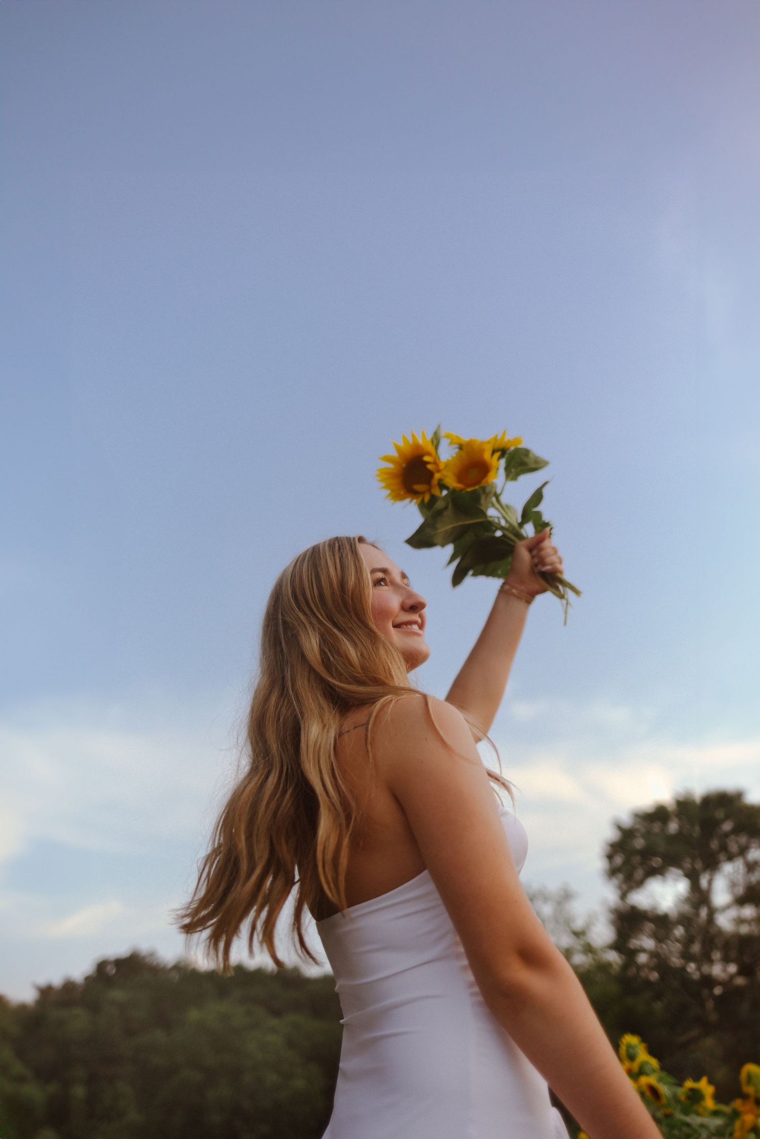 Person in white dress stands in a sunflower field at sunset, holding sunflowers up to the sky with flowing hair.
