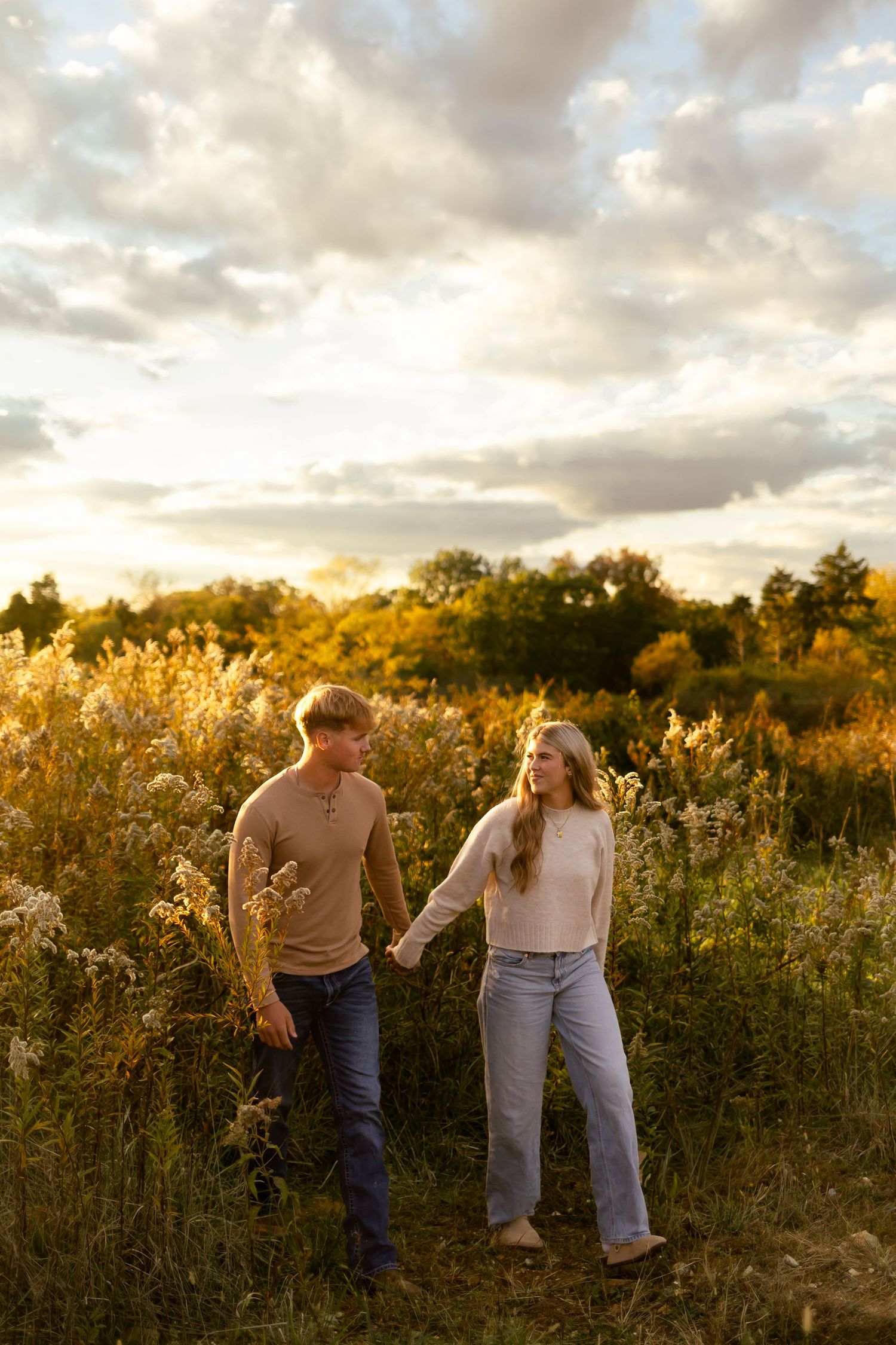 A couple walks hand in hand through a golden meadow at sunset with dramatic clouds overhead.
