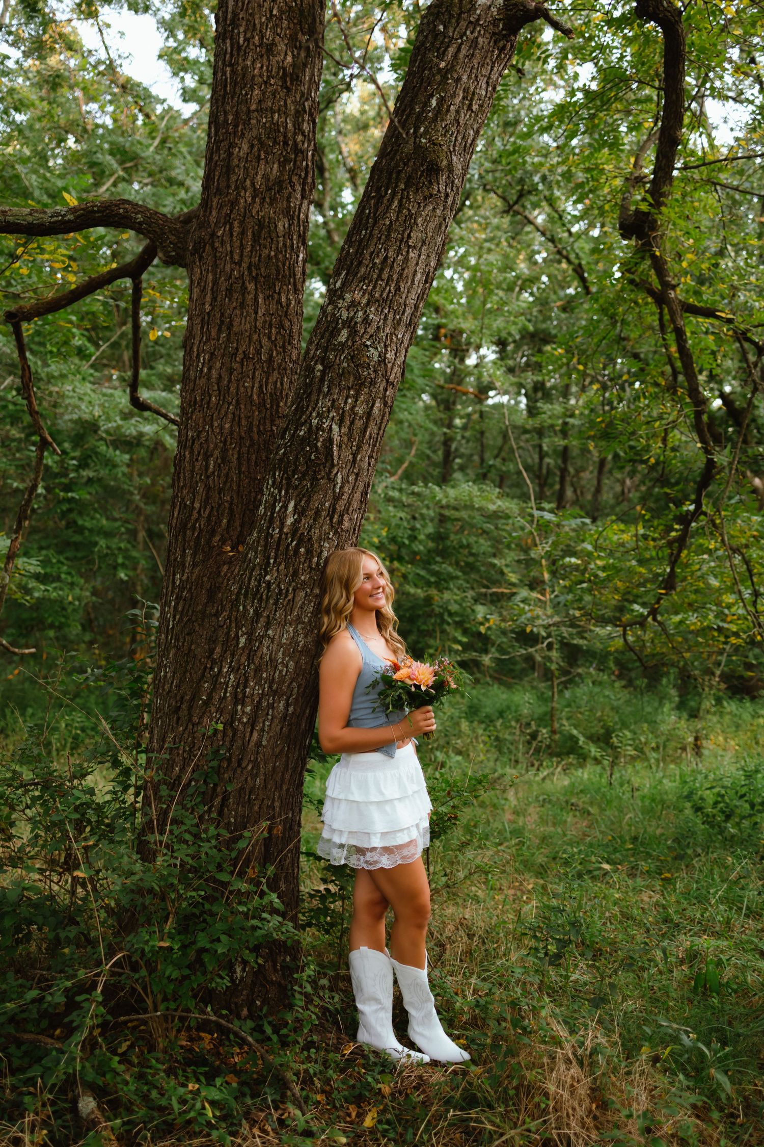 Young woman in white dress and cowboy boots holding orange flowers, leaning against a large tree in a lush green forest.