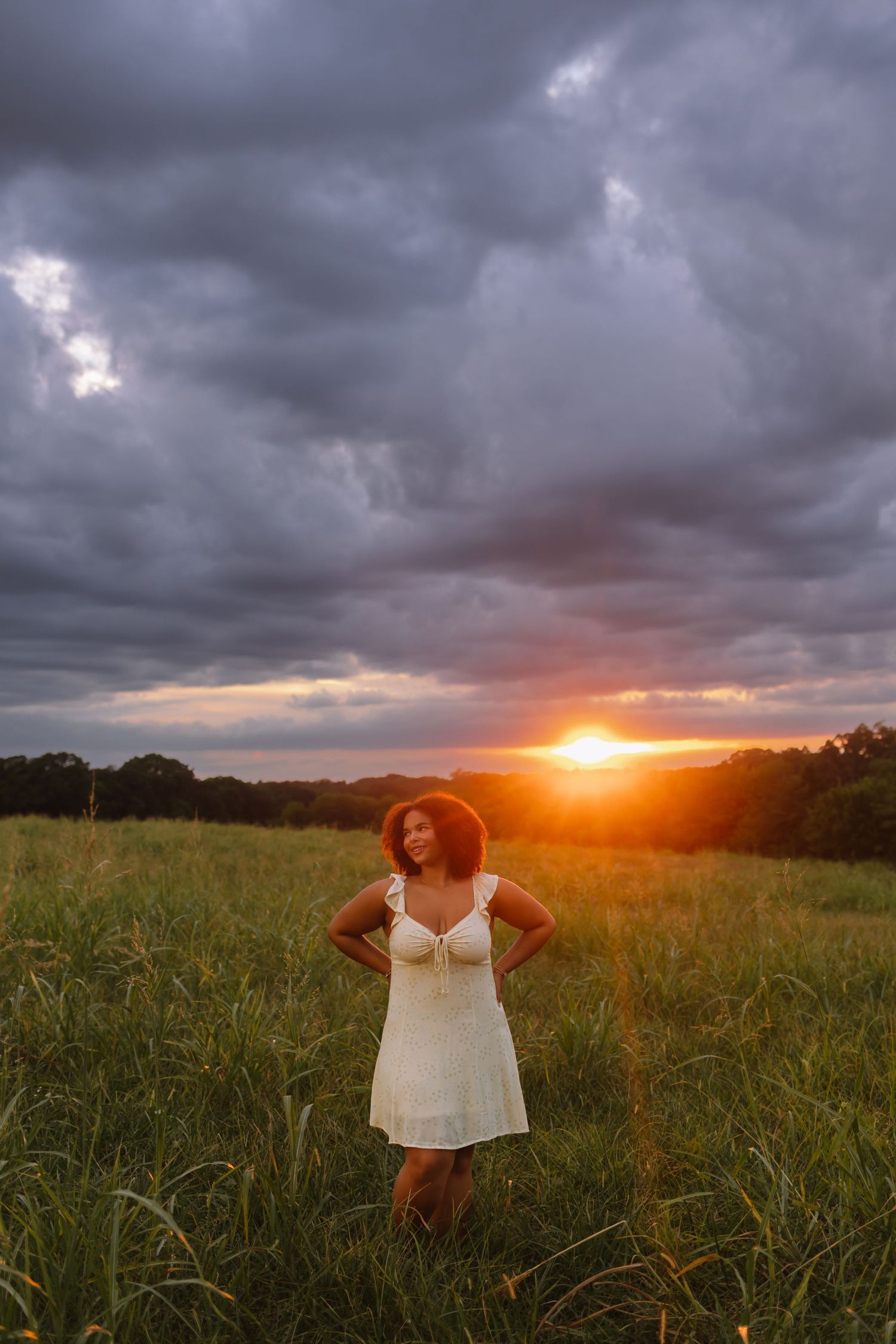 A silhouette stands in white dress against dramatic stormy clouds and golden sunset in a grassy meadow.
