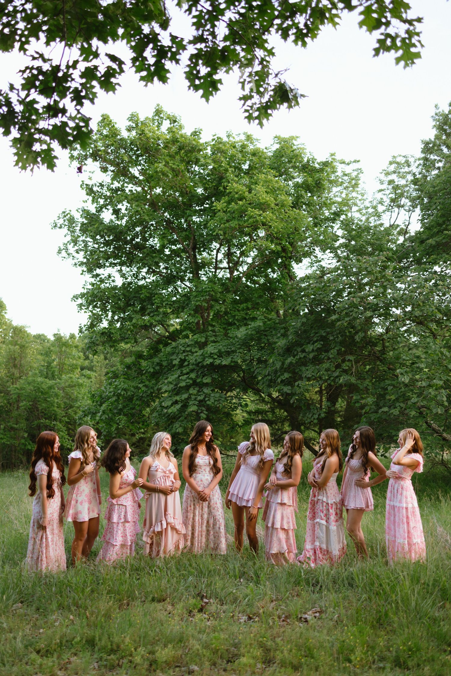 Group of bridesmaids in mismatched pink dresses standing in a circle outdoors surrounded by lush green trees.