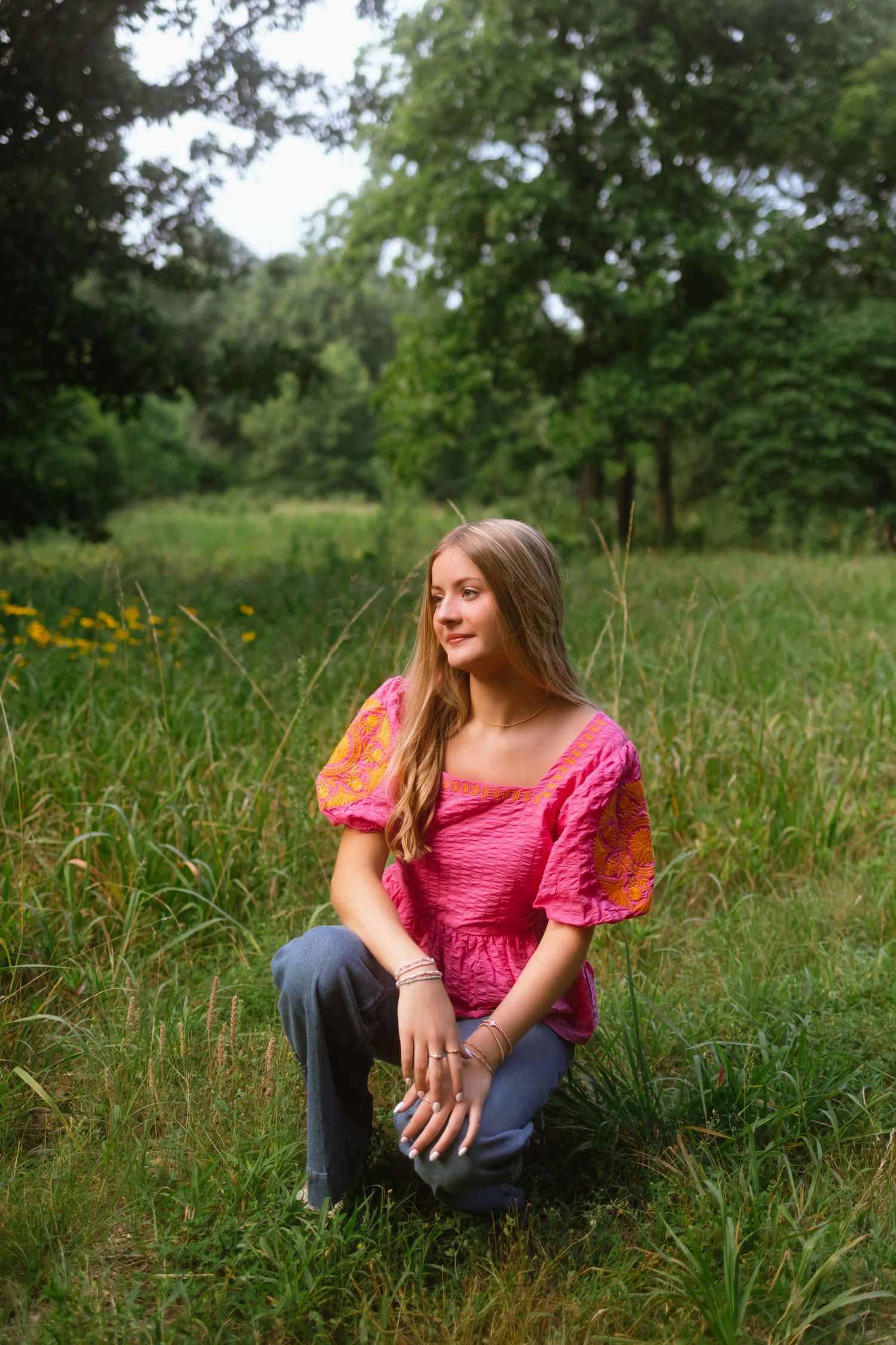 Young woman in pink top and gray jeans kneeling in a lush green meadow surrounded by trees.