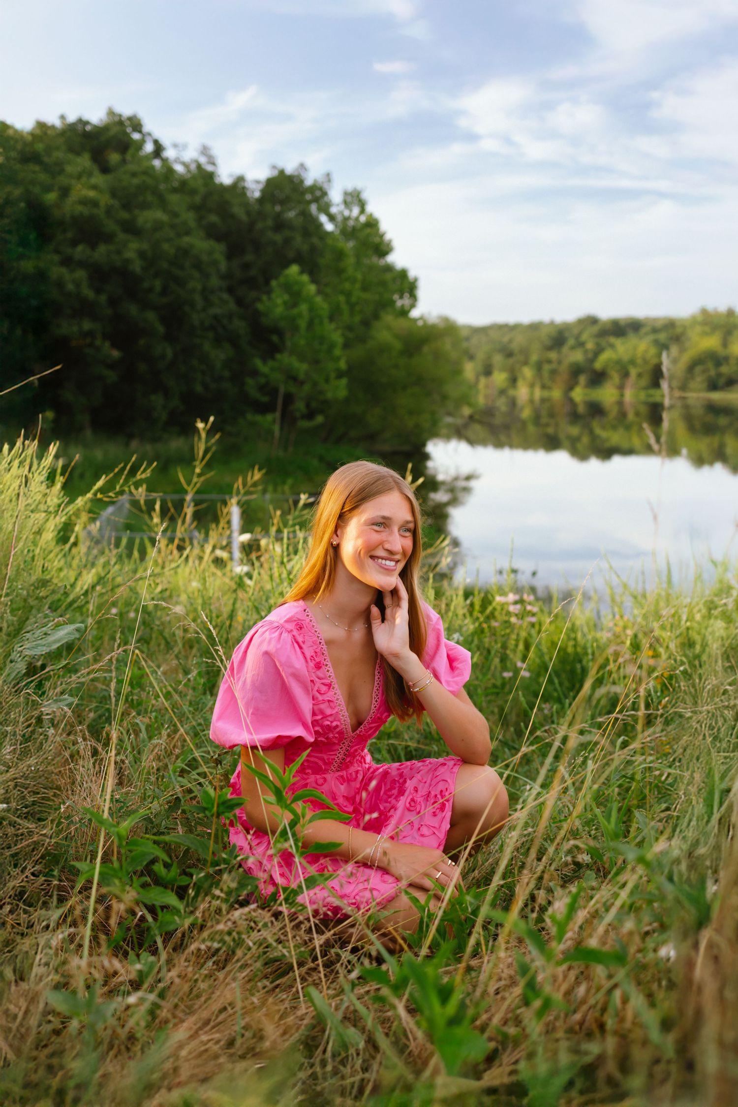 Smiling young woman in pink dress sitting in grass by a serene lake surrounded by lush green trees.