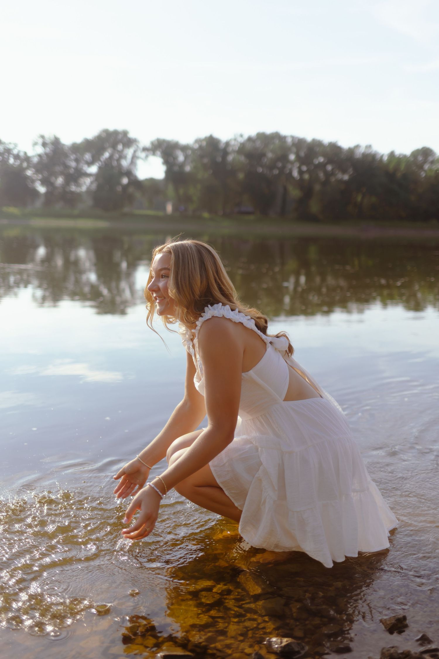 A person in a white dress sits by a lake at sunset reaching into the sparkling water creating ripples.
