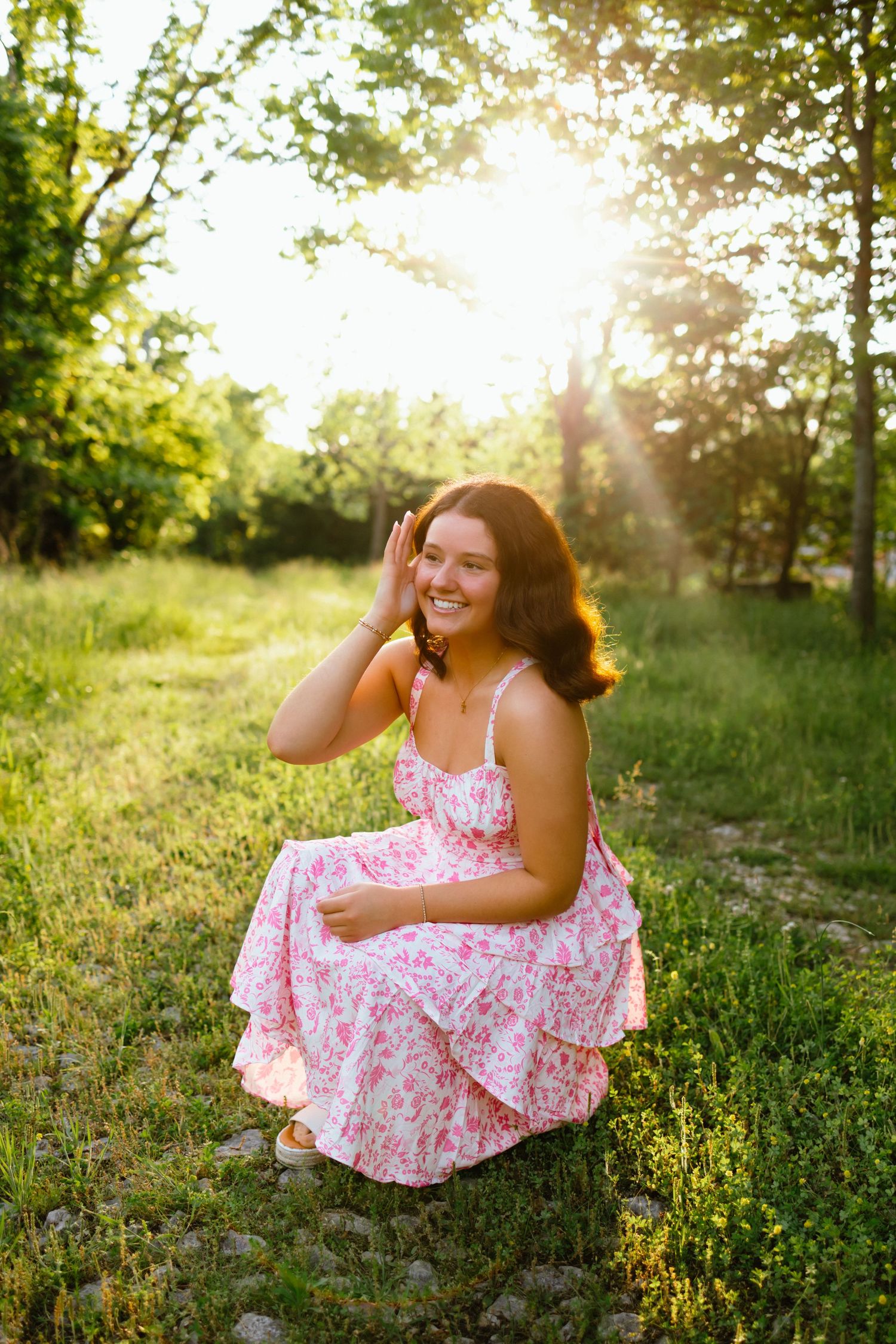 Young woman in pink floral dress crouching in sunlit green meadow, smiling with hand raised near face.